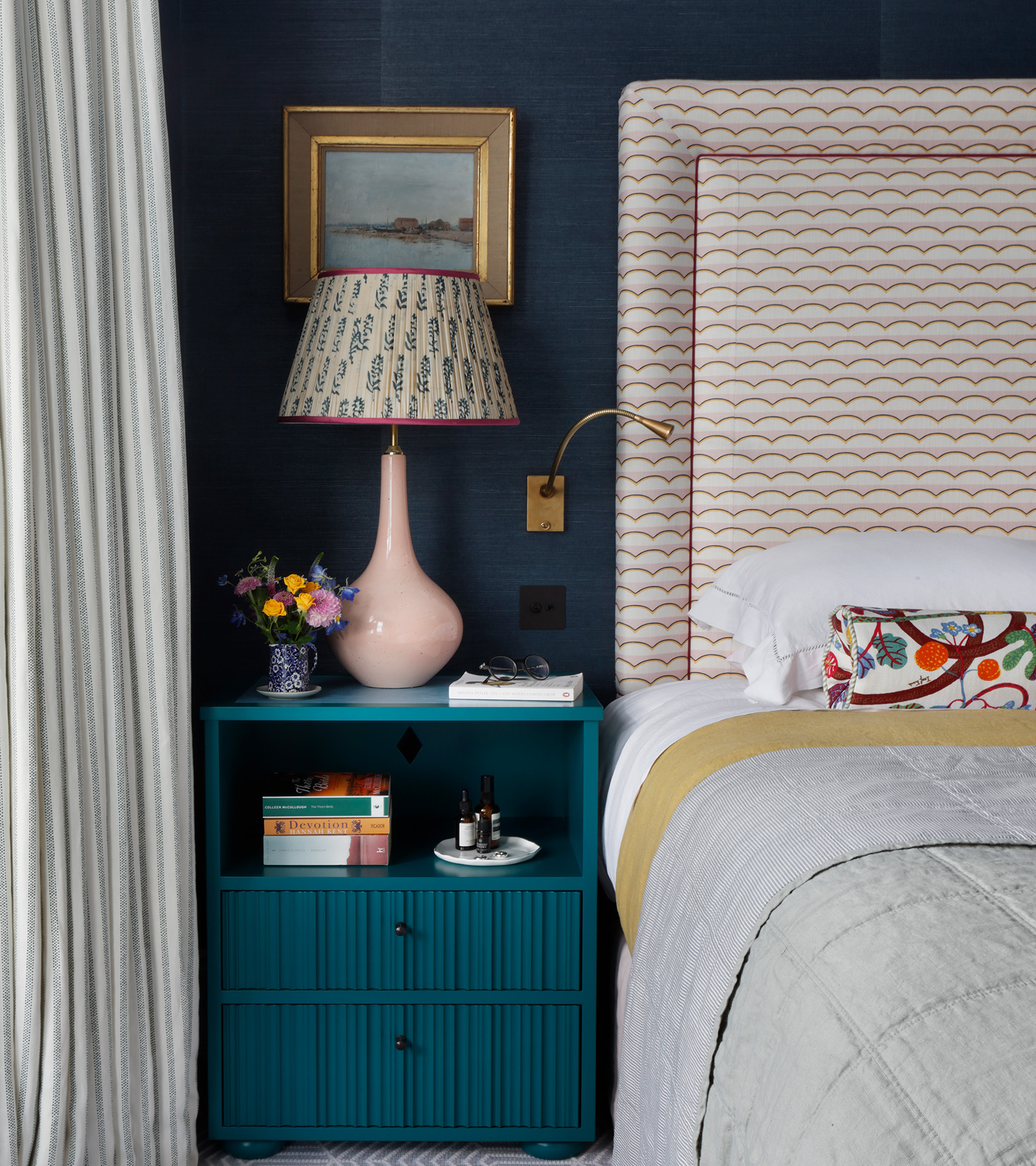 a bedroom with dark navy walls, a teal blue bedside table with a large pleated lamp on top, a bed with white bedding and a pink and white patterned headboard