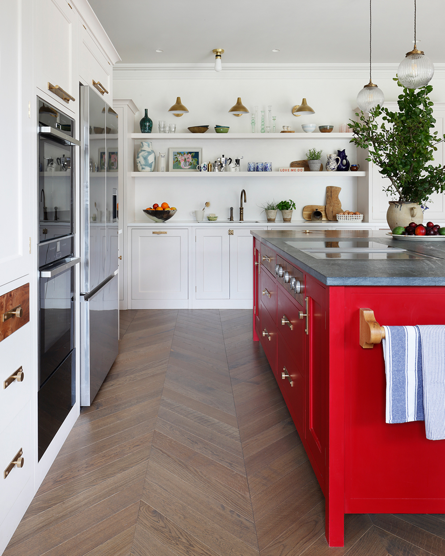 a white kitchen with a bright red painted kitchen island with a dark grey marble top, a vase with leaves and 2 glass ball pendant lights above it