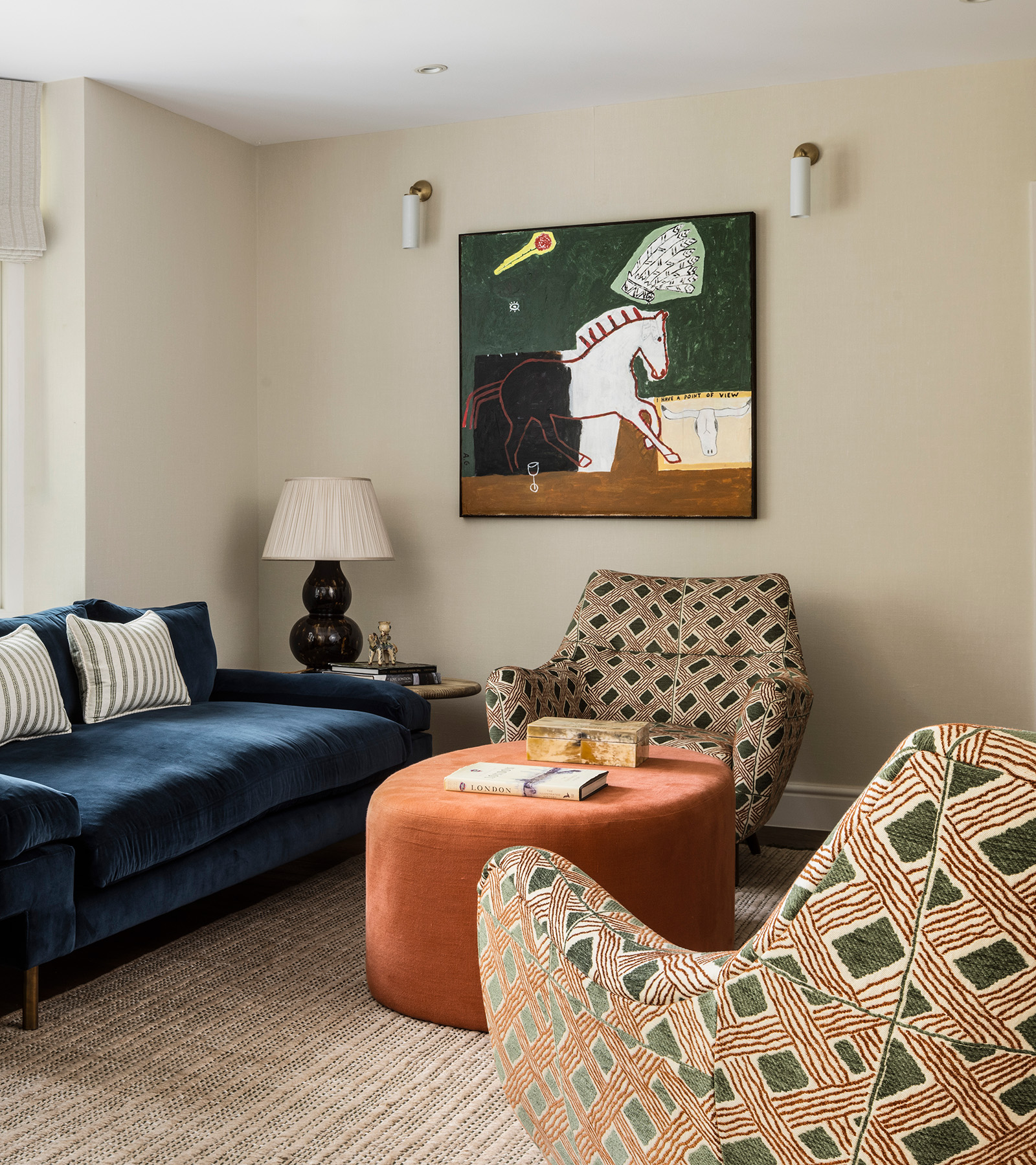 a living room corner with neutral walls and a large art print of a horse on the wall with two lights next to it, a blue sofa and two patterned accent chairs with a round orange ottoman