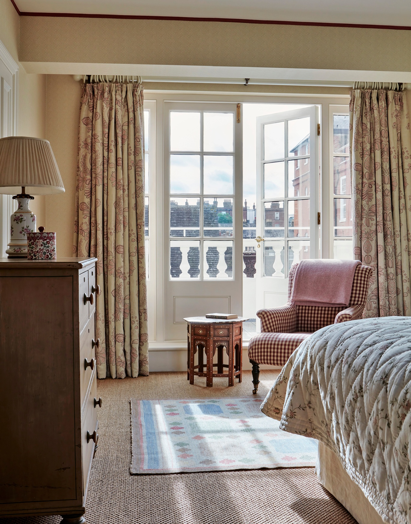 A shot of a bedroom with a check armchair and antique table in front of half-open French doors opening out onto a balcony in the background. In the foreground to the left is a chest of drawers and to the right is the end of the bed dressed with a quilt