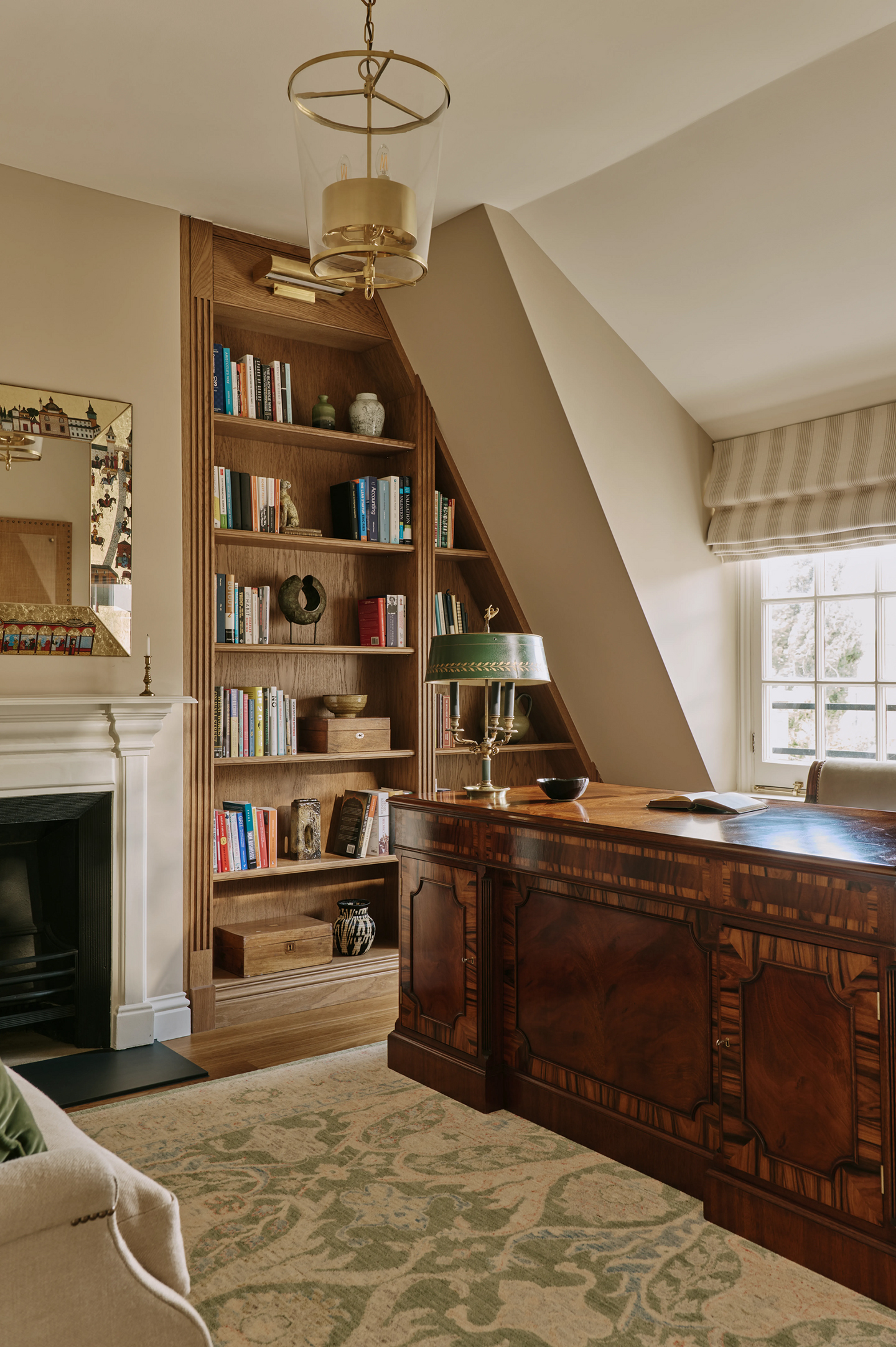 A home office with a large wooden desk, topped with a traditional table lamp and stationery. A large rug covers most of the wooden floor, and a wooden bookcase has been built in next to the fireplace a pendant lantern hangs above