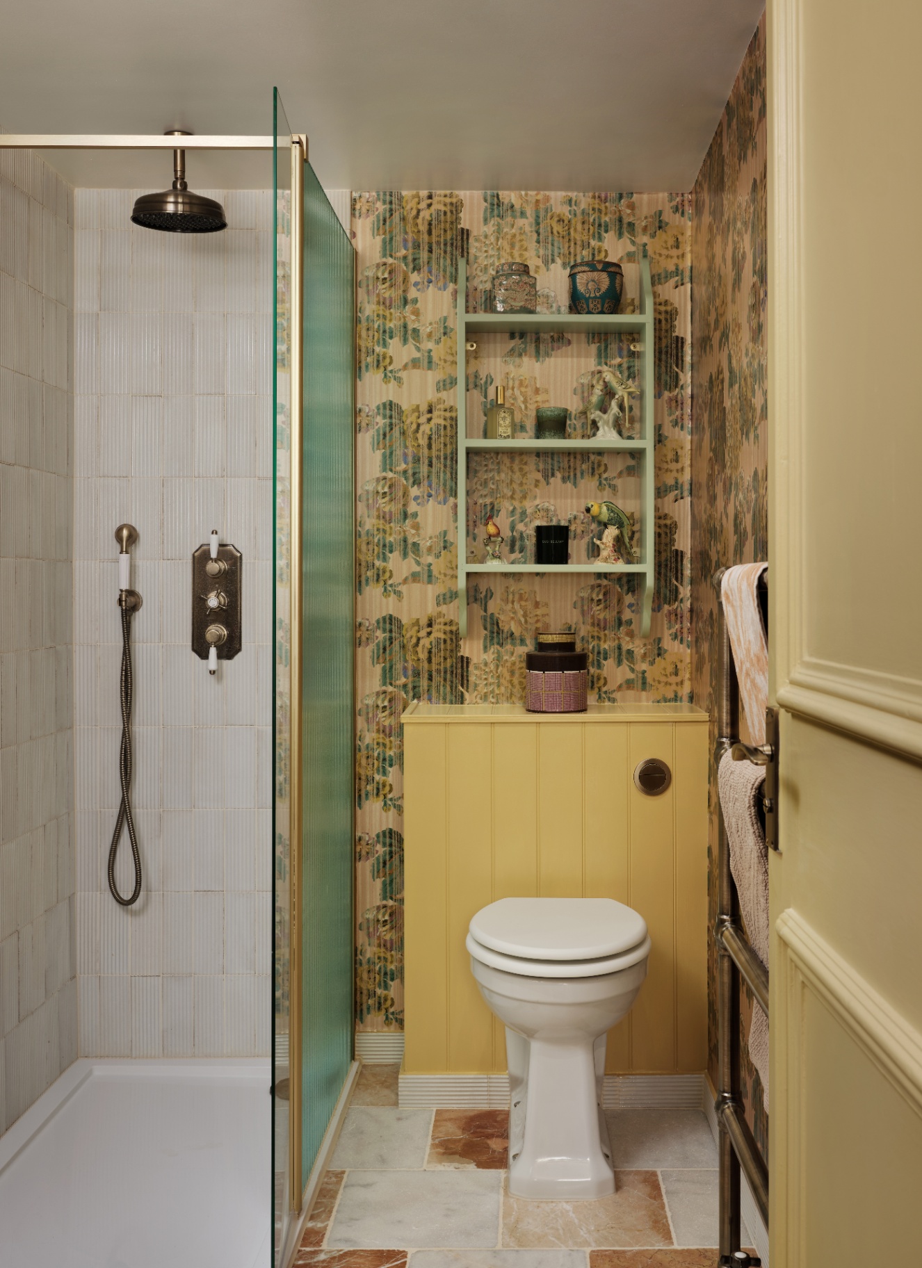 a small bathroom witha shower cubicle, gold hardware, yellow panelling behind the toilet and floral wallpaper