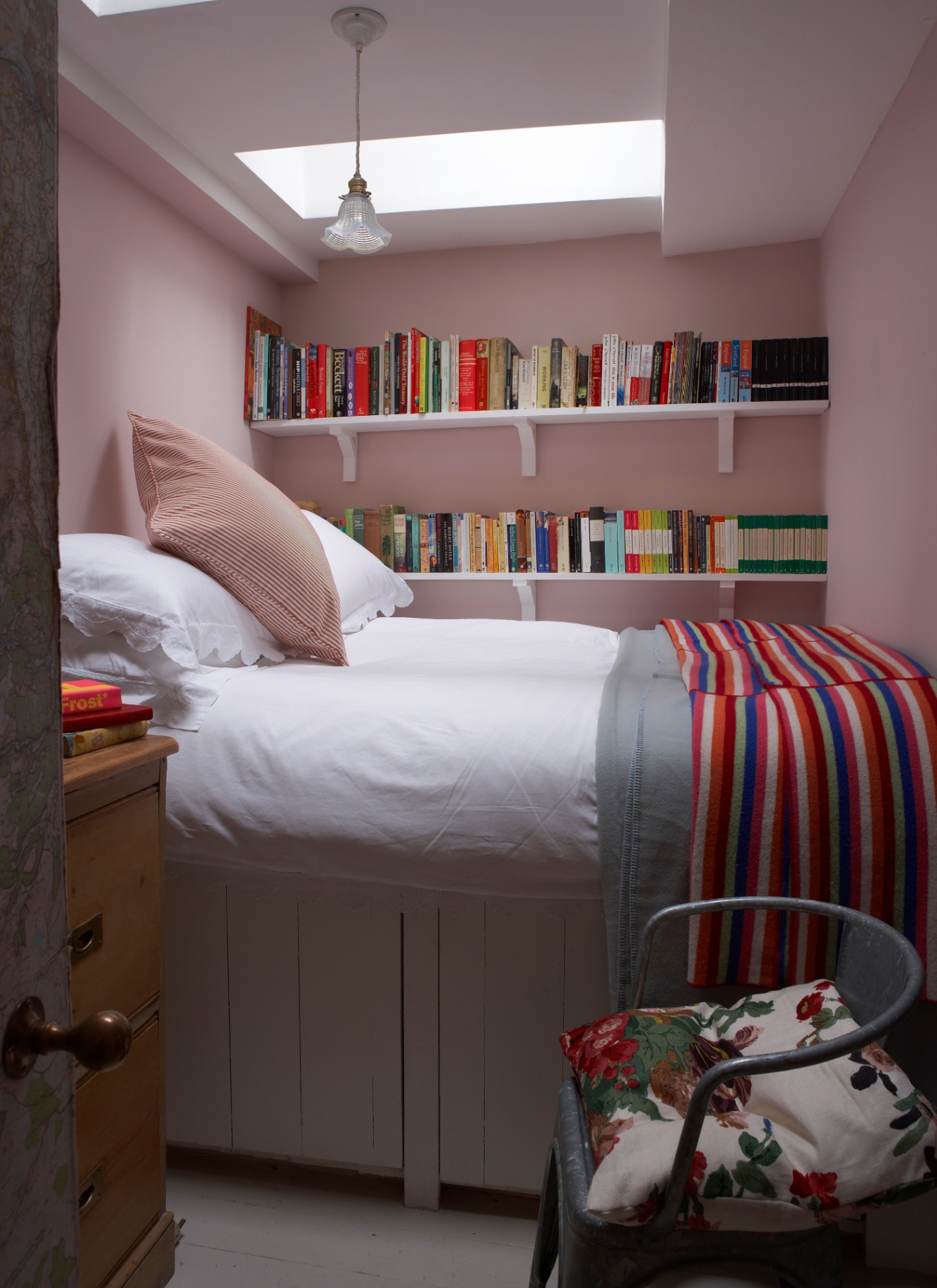 a small bedroom with pale pink walls and two rows of shelves on the back wall above the bed filled with colourful books