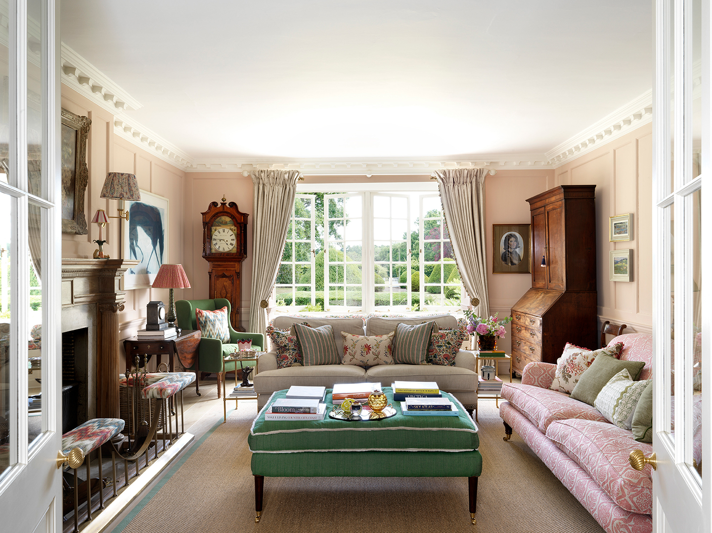 A pale pink living room with a green ottoman in the centre, surrounded by a cream sofa, a patterned pink sofa and a fireplace with a flamestitch fender. French windows open to the garden in the background, with antique brown furniture either side