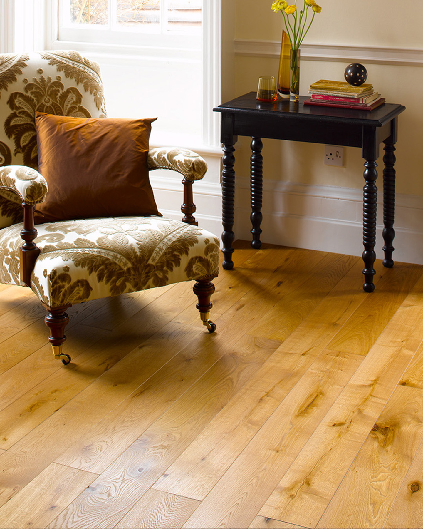a close up of wooden flooring with a white and brown patterned upholstered arm chair with a rust orange cushion and a black console table with a stack of books and a vase with a bunch of yellow flowers
