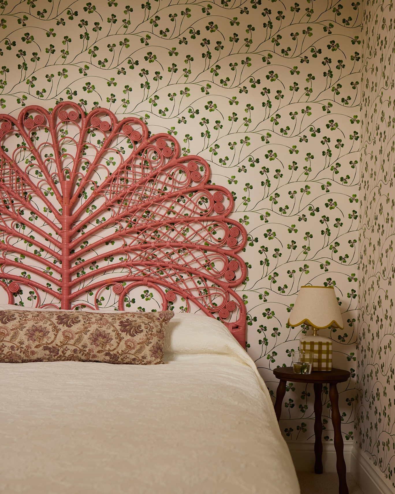 A close up of a bedroom with a clover wallpaper covering the walls. There is an intricate pink rattan headboard and a small wooden side table topped with a table lamp and glass of water. A decorative floral cushion is sat on top of the bed 