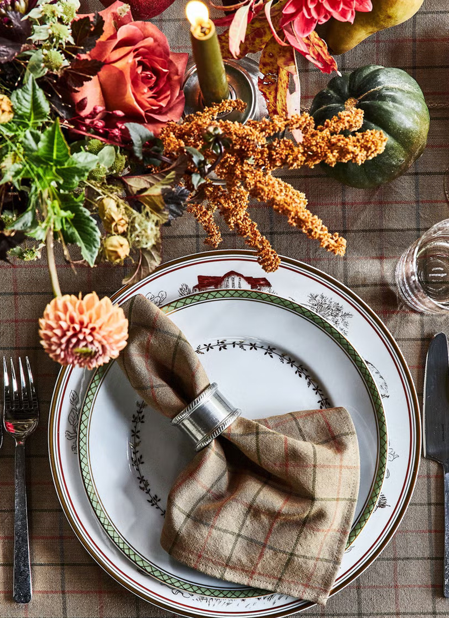 a close up of an autumn table with a beige, dark green and orange checkered tablecloth and matching naokin on a white plate with a silver napkin ring, silver cutlery, an autumnal floral arrangement and a green dinner candle