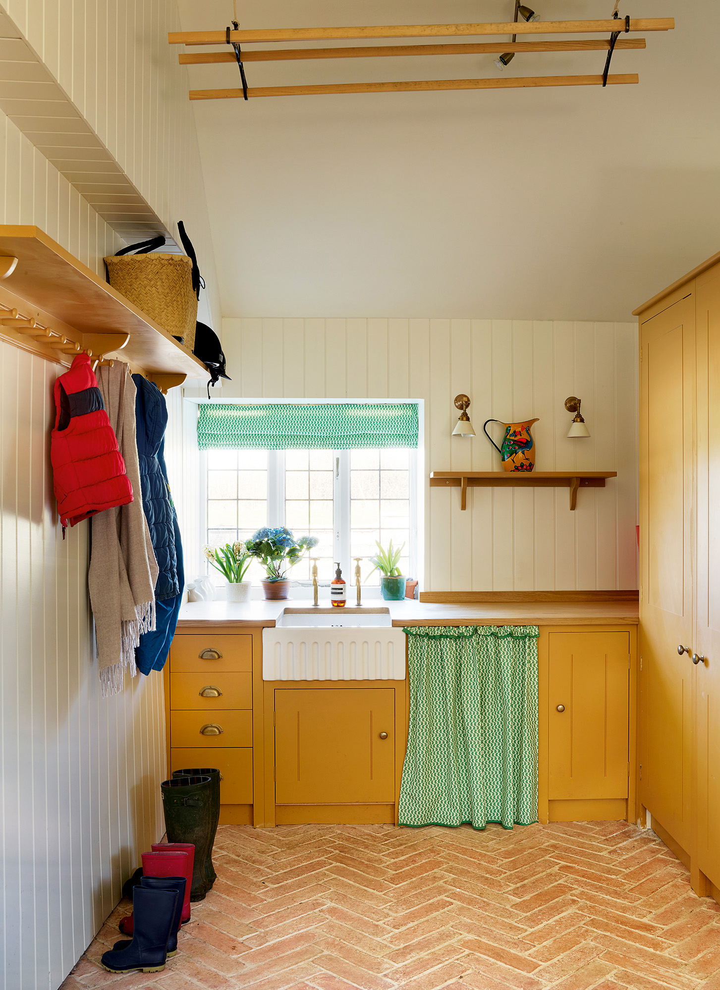 a utility room with mustard yellow cabinets, a belfast sink, a green and white patterned blind and matching sink skirt and brick flooring
