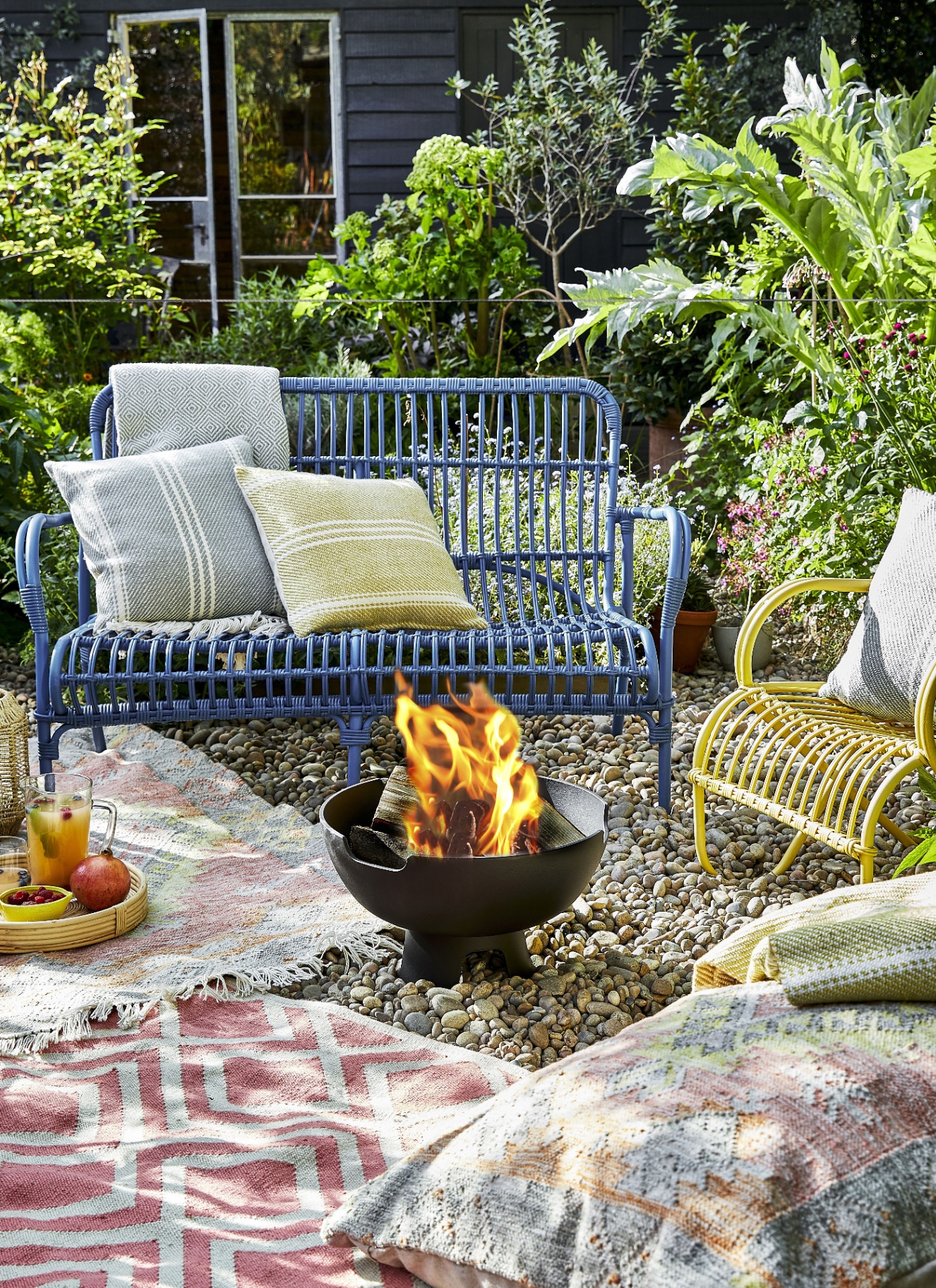 a garden with a metal blue bench and a metal yellow chair with cushions and a fire pit in front