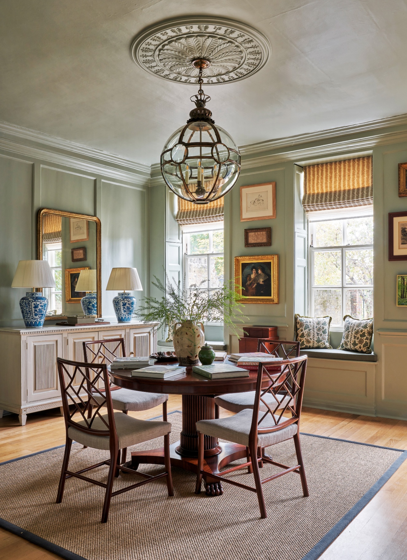 Sage green panelled dining room with a round wooden table, lattice-back dining chairs, globe pendant light, blue glazed lamps, and woven sisal rug on wooden floors.