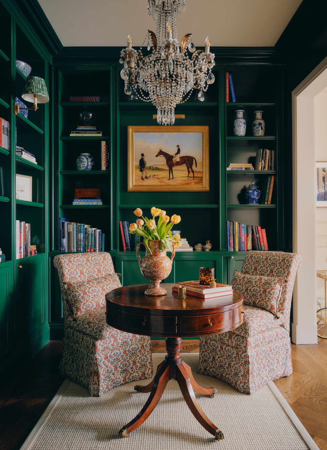 a library corner with bookshelves painted in emerald green, a large crystal chandellier hanging above a round antique table with two white and orange floral slipper chairs