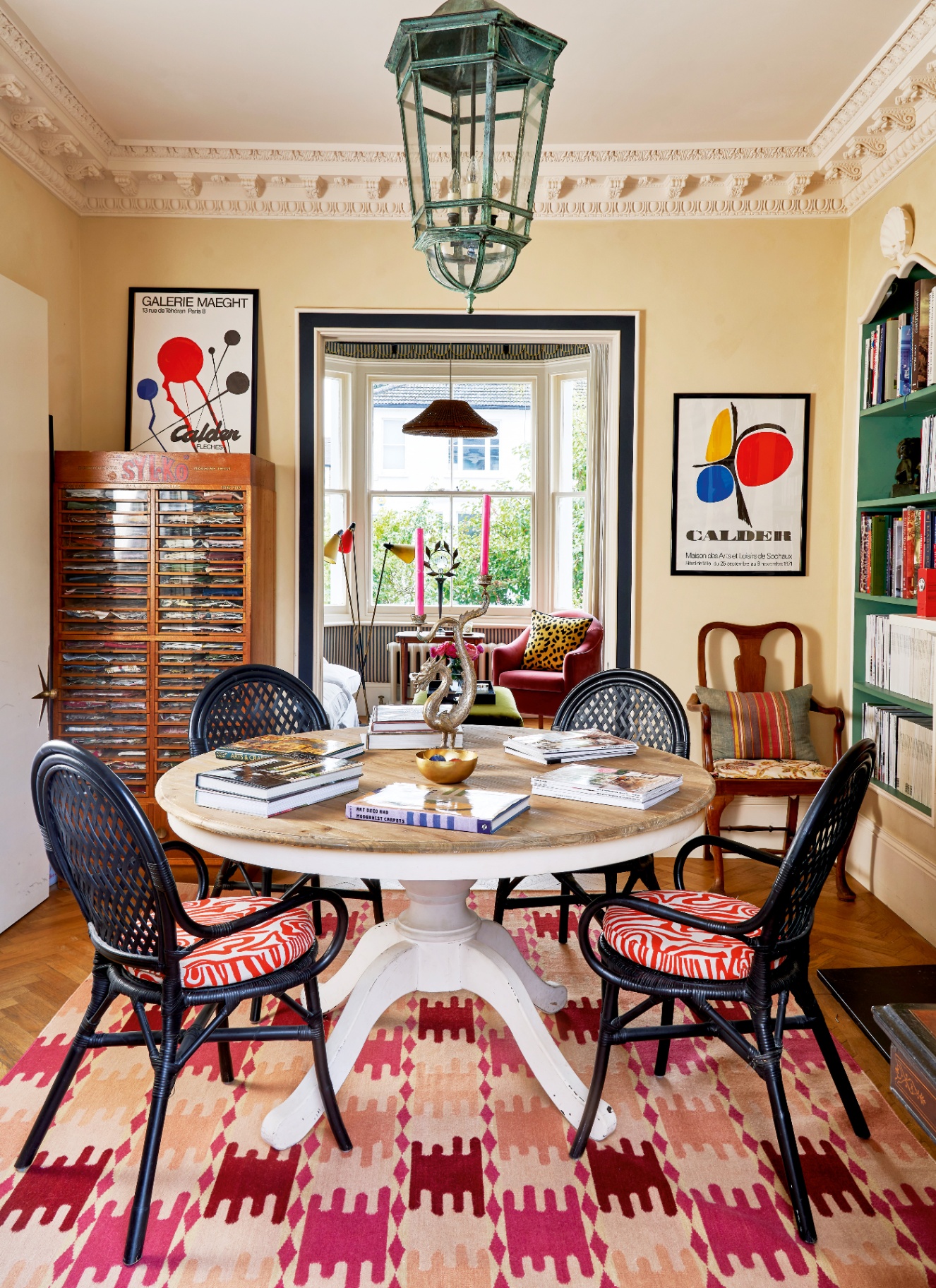 Eclectic dining library with a round pedestal table, black woven chairs on a pink patterned rug, framed art, and green bookcases