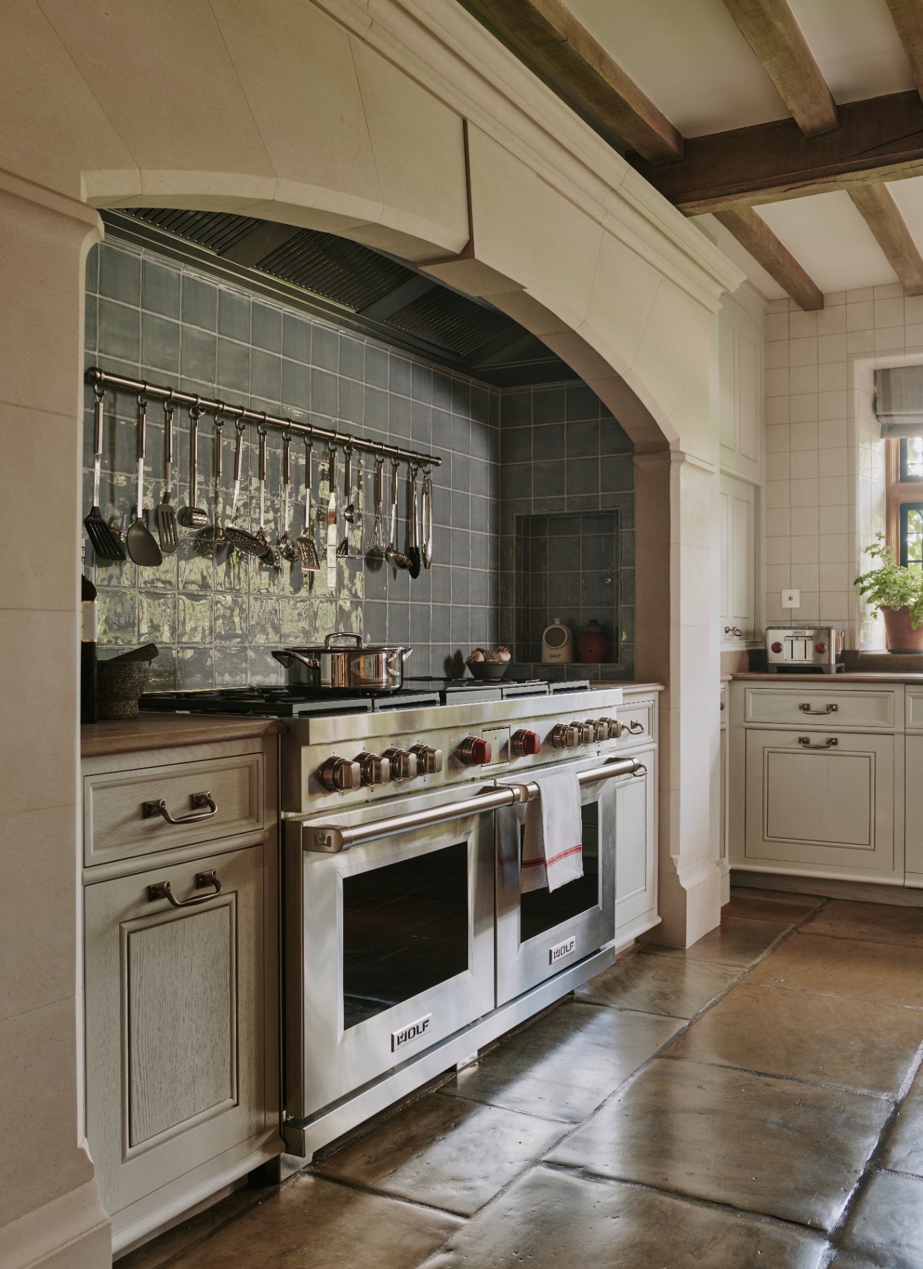 an english kitchen with white walls, stone floors, a large silver range cooker and an alcove covered in dark blue tiles