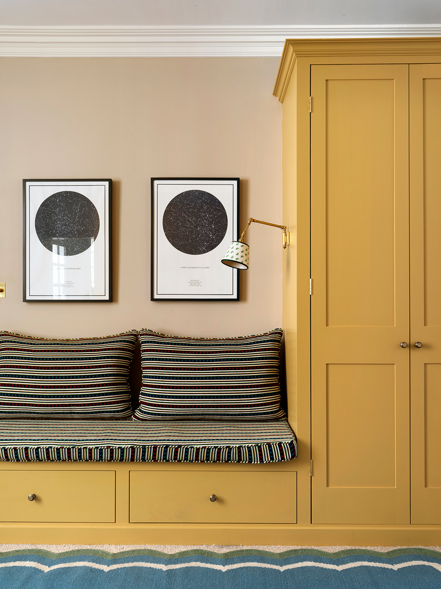 A close up of a playroom with built-in yellow joinery including a tall cupboard and drawers topped with striped cushions to create a bench seat. Two celestial prints hang on the wall and the edge of a scalloped blue and green rug can be seen