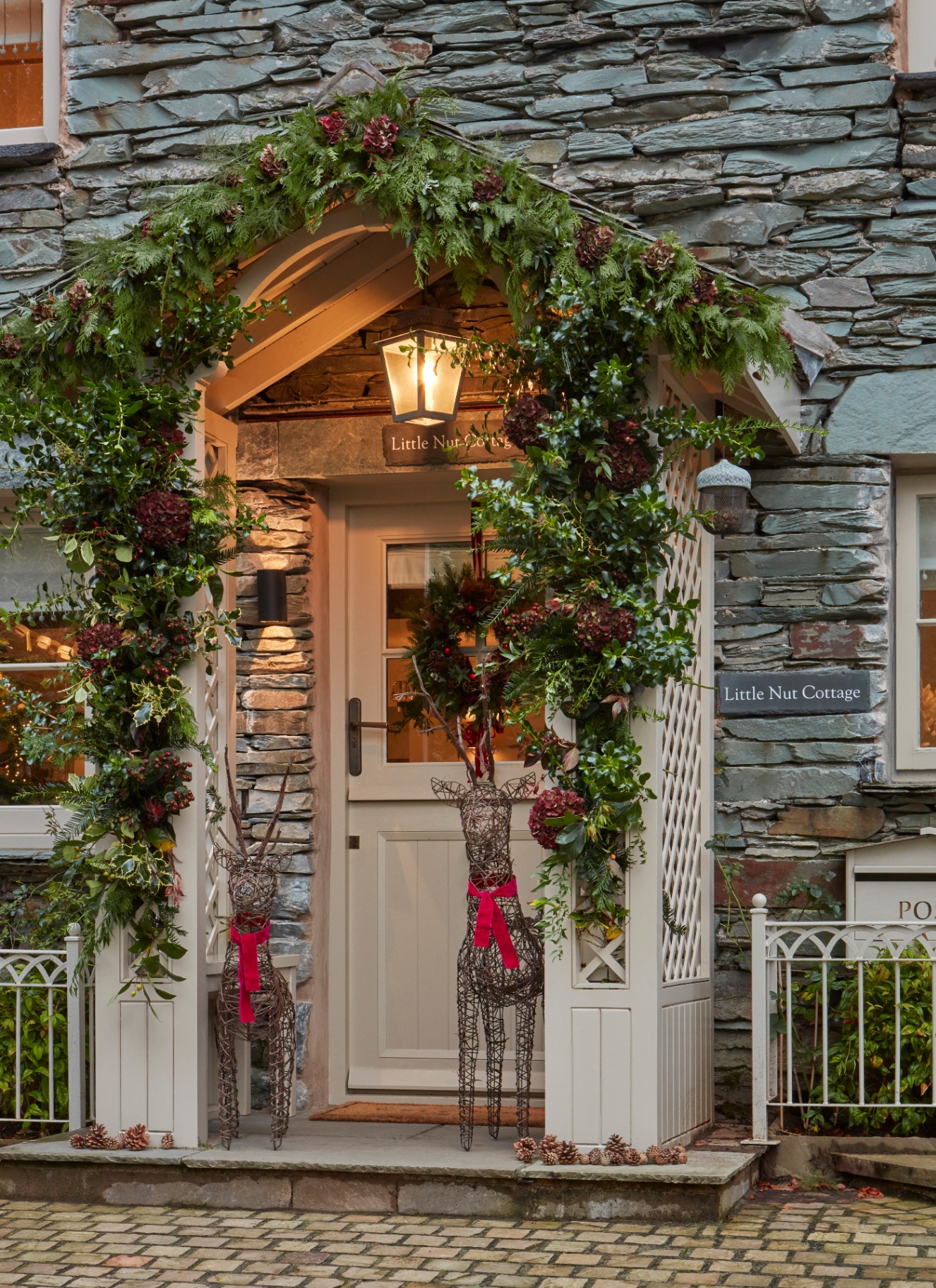 Two woven wicker reindeer with red scarves standing on a stone porch flanked by a dramatic foliage archway over the front door