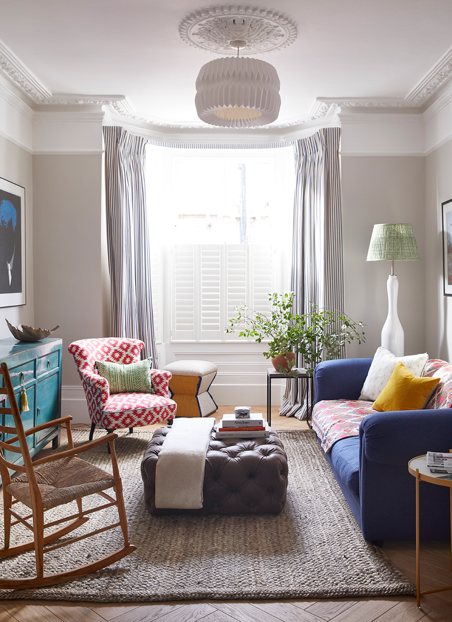 Small living room with a blue sofa, red geometric armchair, and wooden rocking chair arranged around a dark leather tufted ottoman on a jute rug, filled with natural light from a bay window