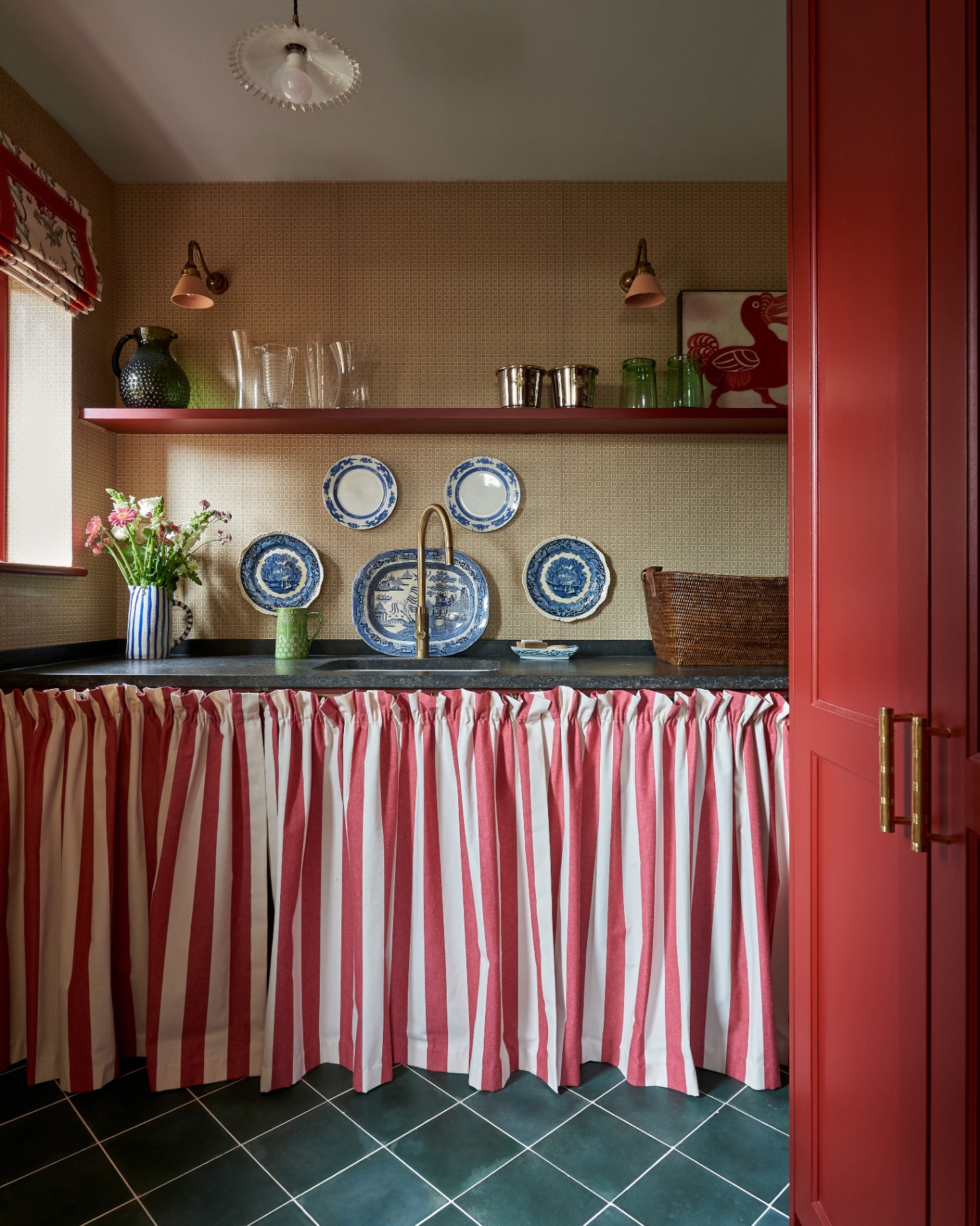 a utility room with a red and white stripe fabric curtain under the sink, ochre and white patterned wallpaper and blue and white patterned plates hanging on the wall