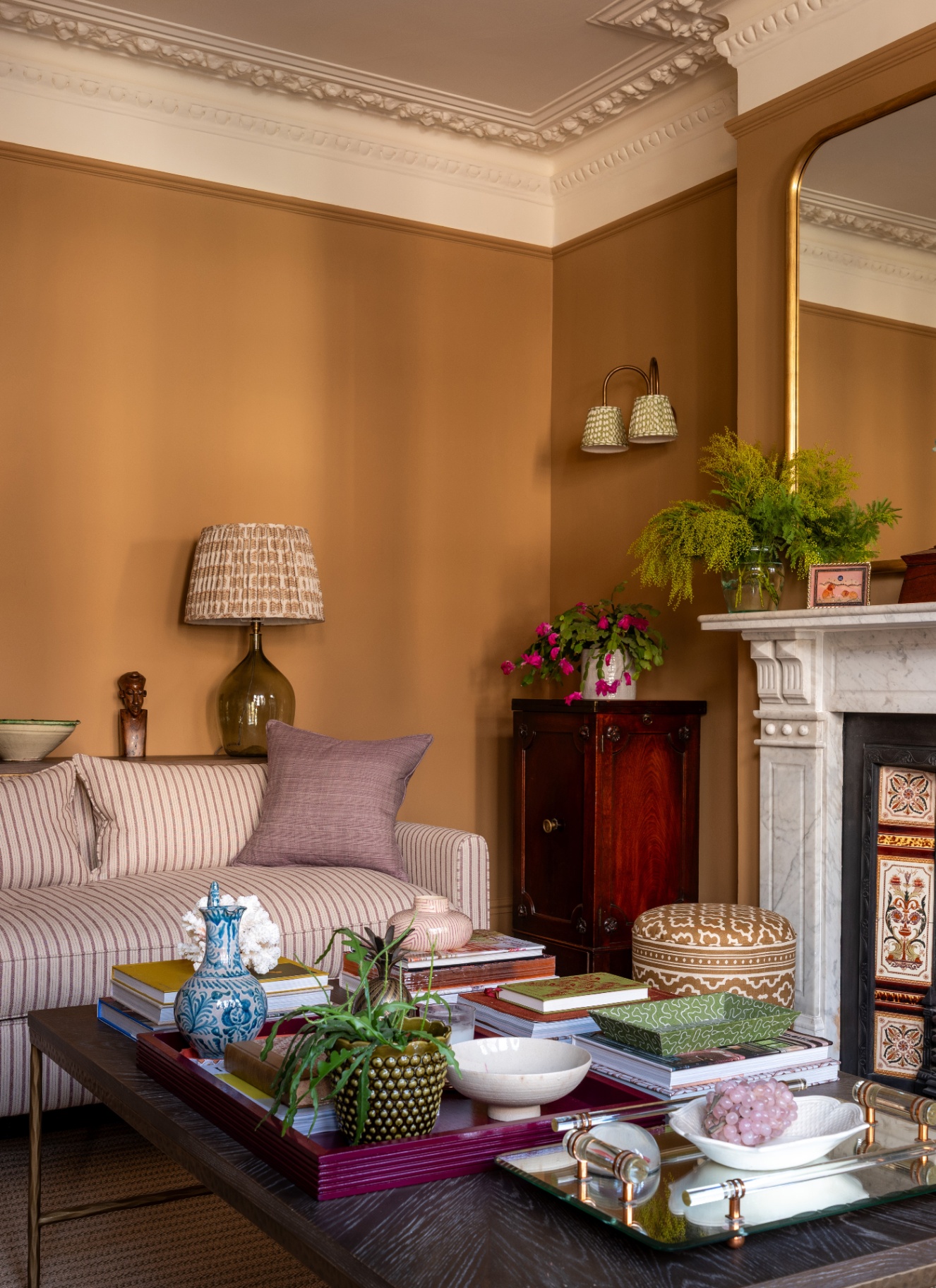 A period living room featuring caramel warm walls, a pinstriped sofa with patterned lamps, and a marble fireplace alongside a coffee table styled with books, plants, and decorative trays