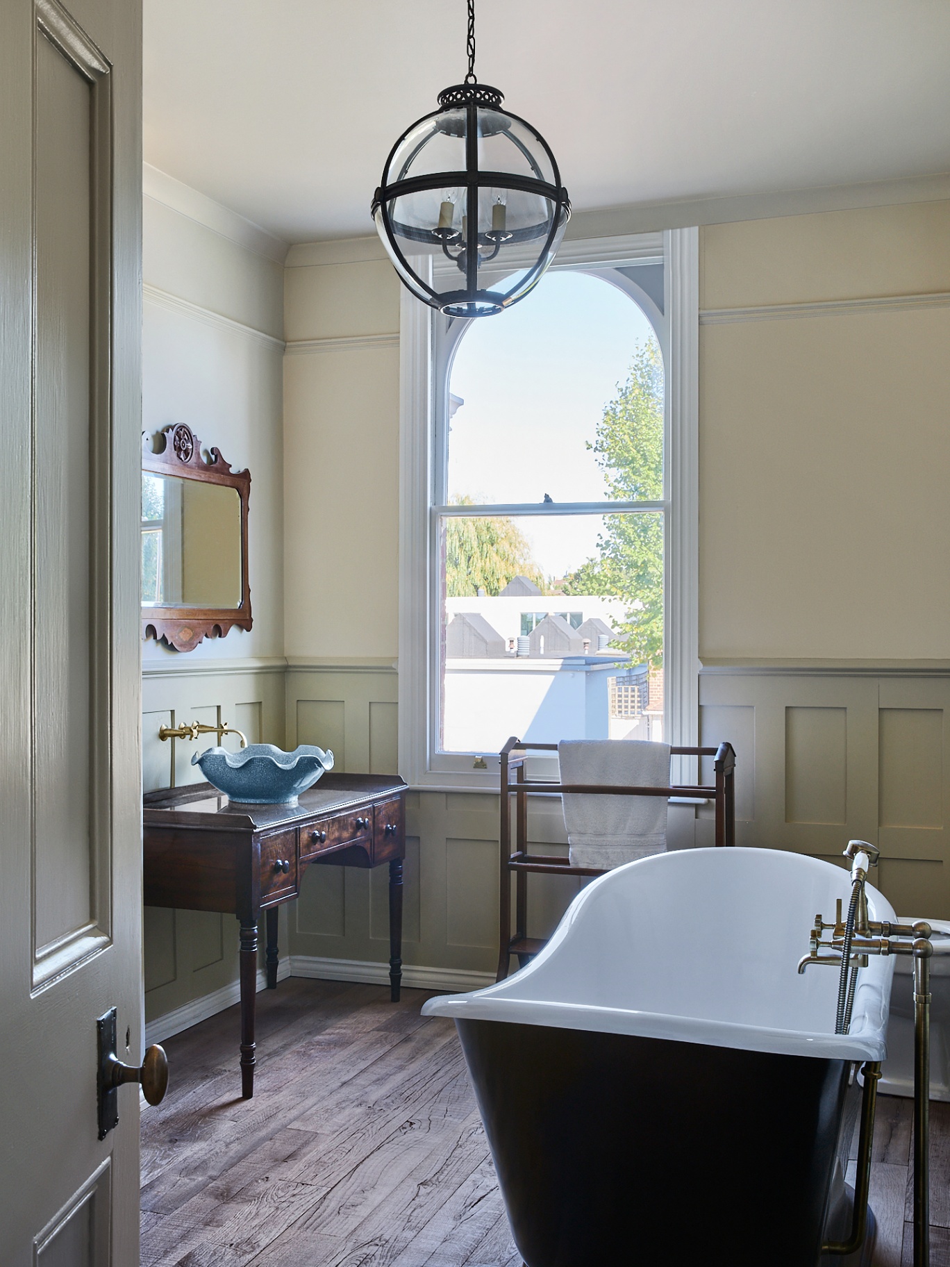 a beautifully lit bathroom featuring antique furniture like a mirror and basin table, on the wooden floors is a roll top bath and and above that a vintage light