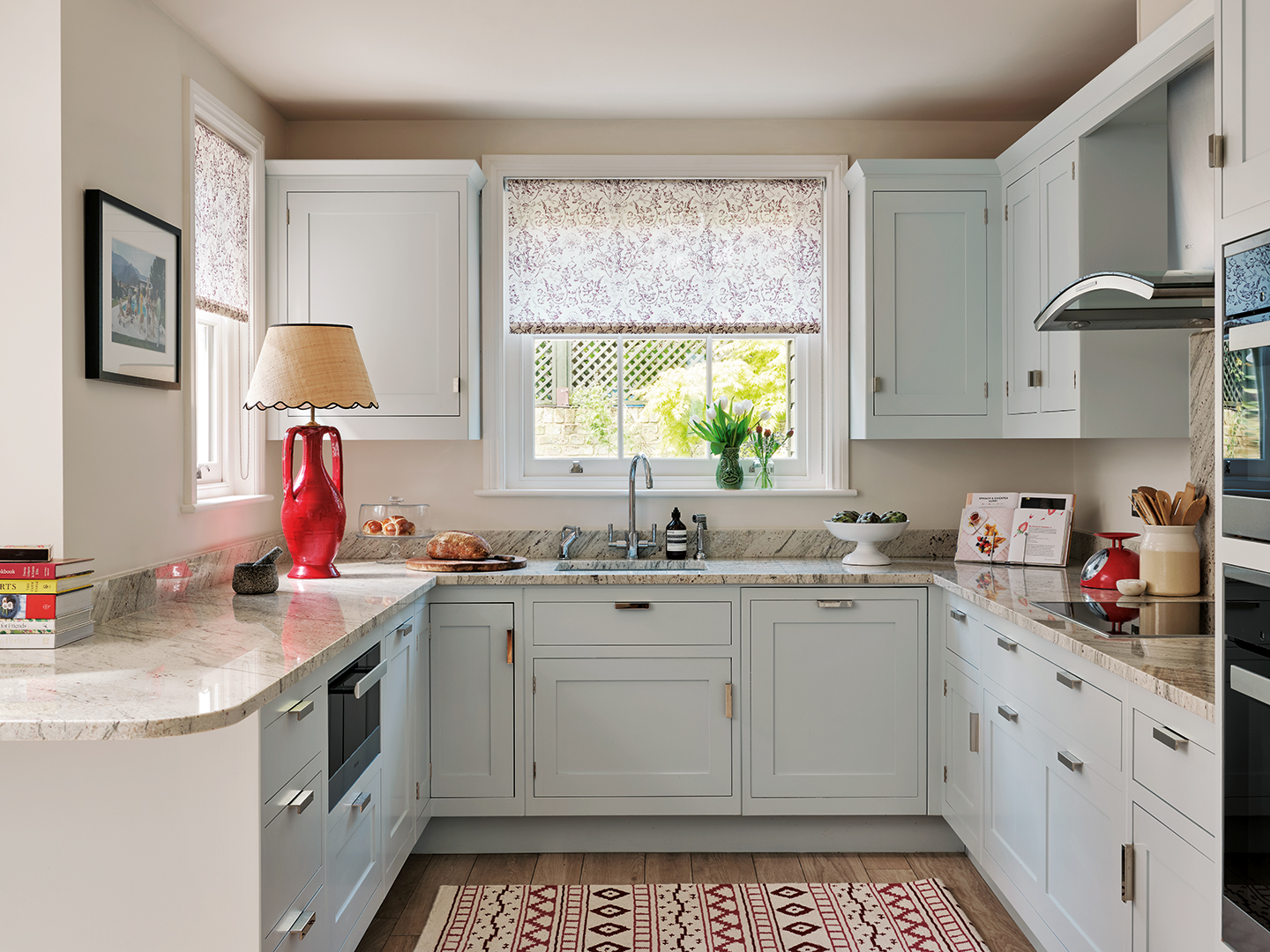 a u-shaped kitchen with pale blue cabinets and marble worktops. A bold red urn lamp base is topped with a rattan shade on the counter and a patterned rug sits on the floor in the middle