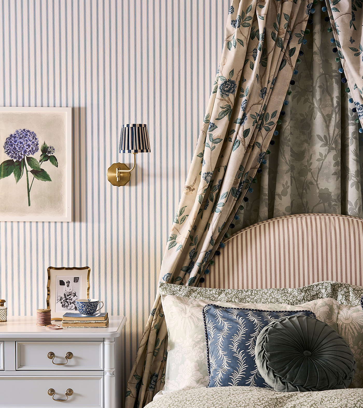 A bedroom with blue and white stripe wallpaper, a framed flower print, a white bedside cabinet, a bed with a floral canopy and a striped headboard