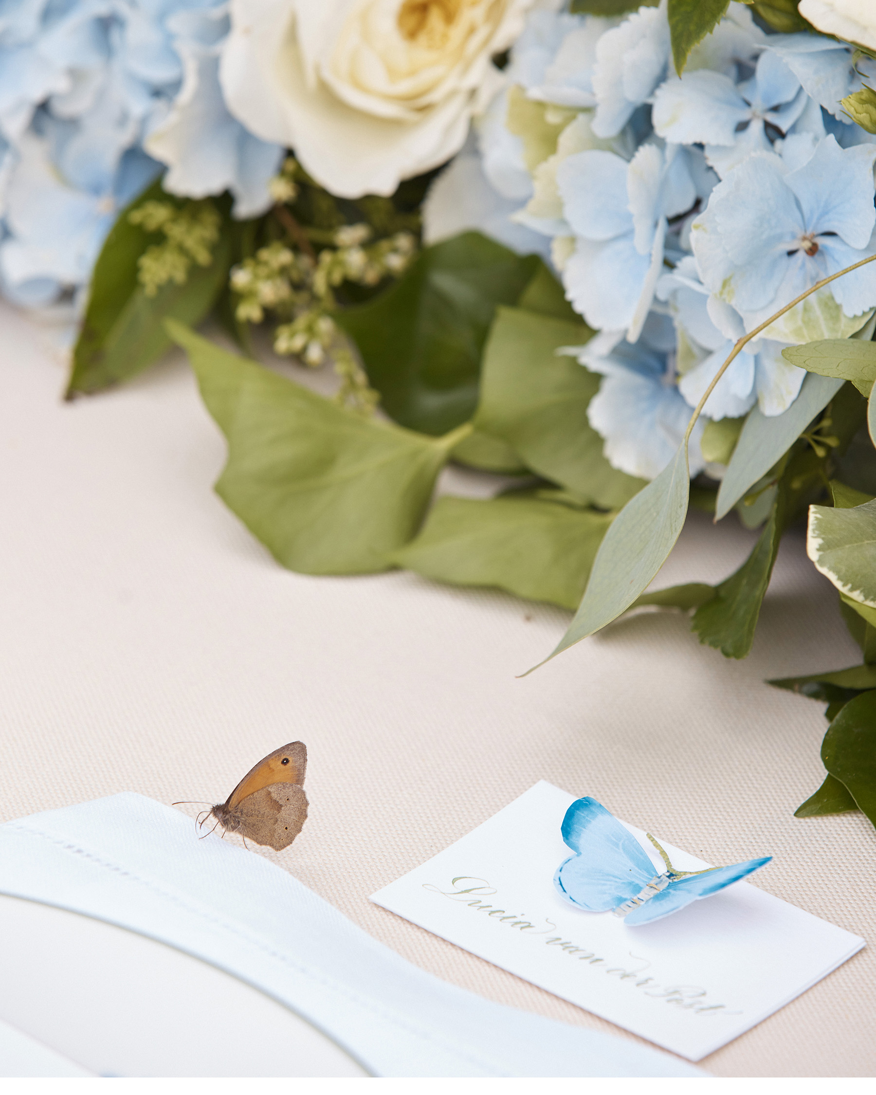 a summer table place name setting with cursive writing and a blue butterfly decoration with a row of blue and white fresh flowers behind it and a real butterfly on the table