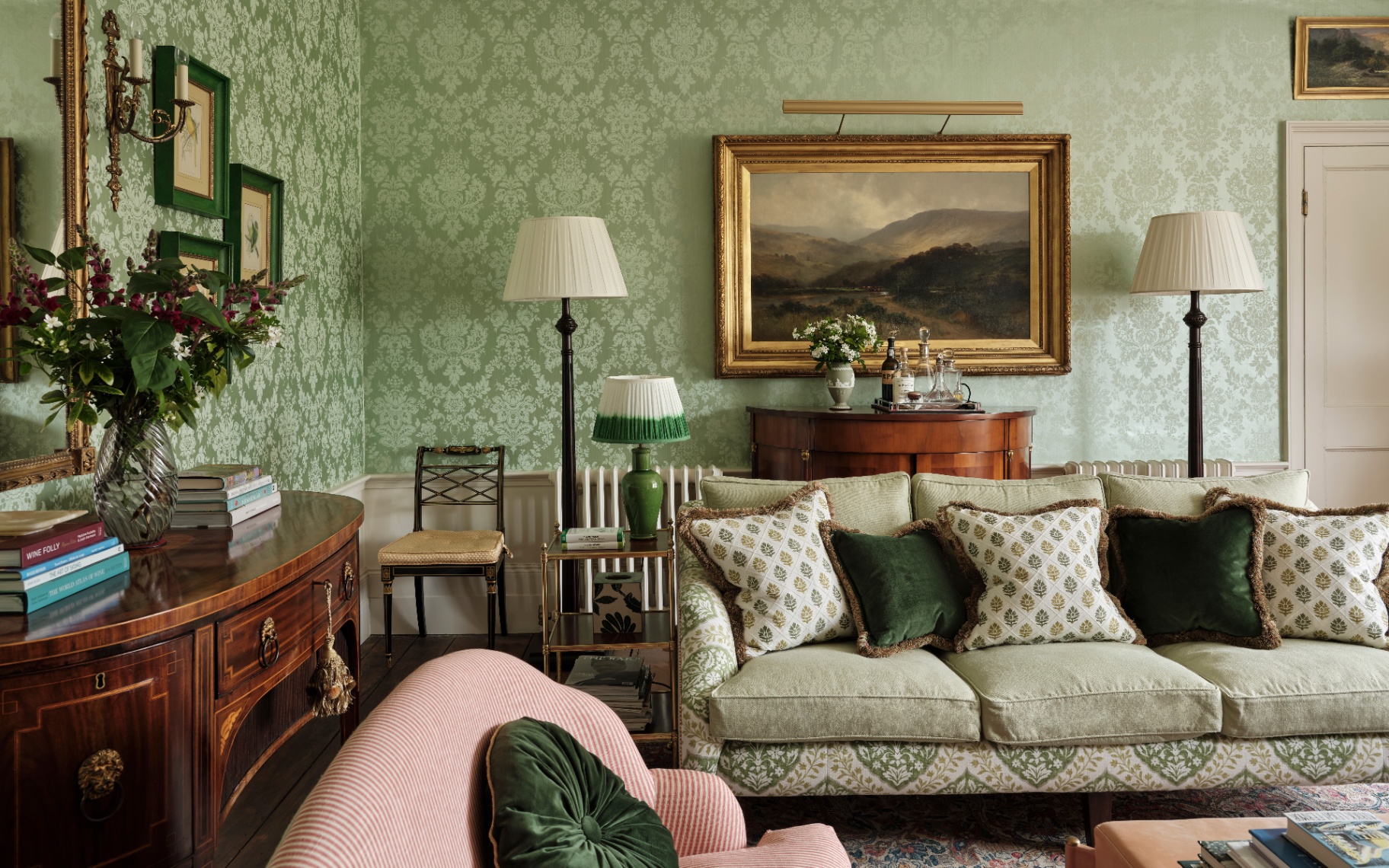 a living room with green floral wallpaper, a white and red stripe sofa, a green floral sofa, a floral rug,a wooden side table and a landscape painting in a gold frame