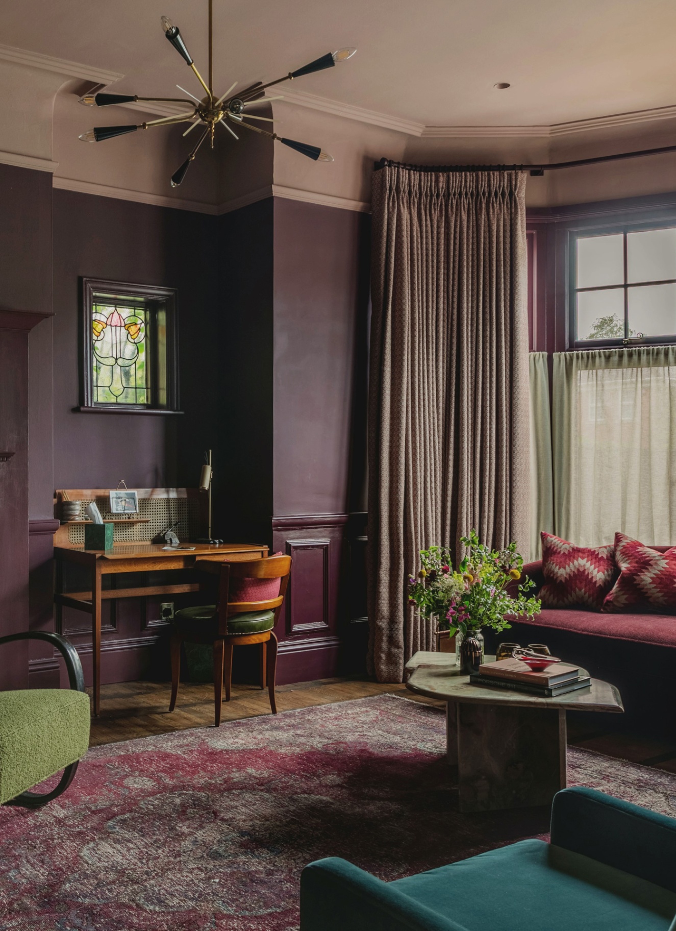 a living room with dark purple walls, beige ceiling, a modern light fitting with six bulbs, a wooden desk and chair against the wall, a red patterned rug and a deep red sofa and long beige curtains