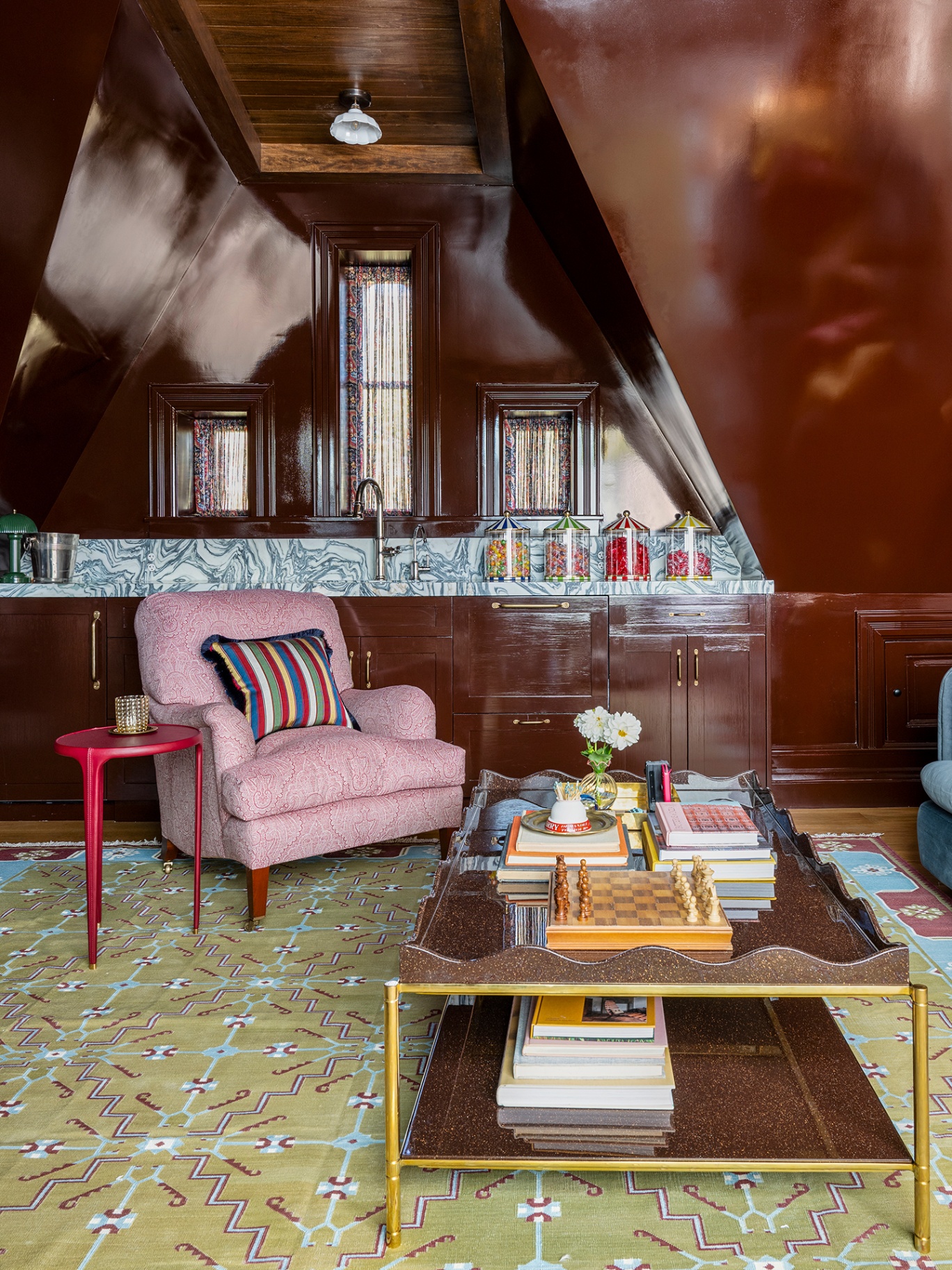 A snug with brown gloss walls, a vibrant yellow-green patterned rug, scalloped coffee table topped with books, flowers and a chess board. An armchair is next to a red tri-leg side table and behind is a marble worktop with a sink and jars of sweets