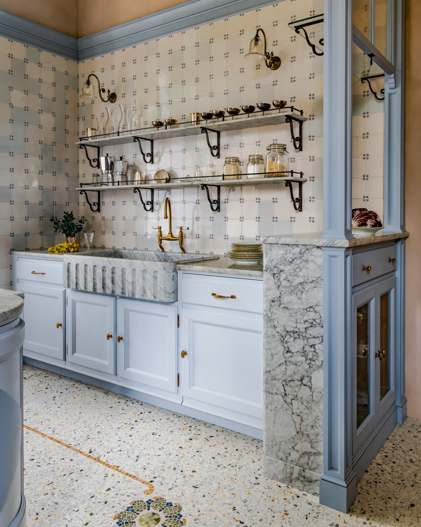 a kitchen with a white terazzo floor, white marble countertops, pale blue joinery and open shelving above the sink with jars on top