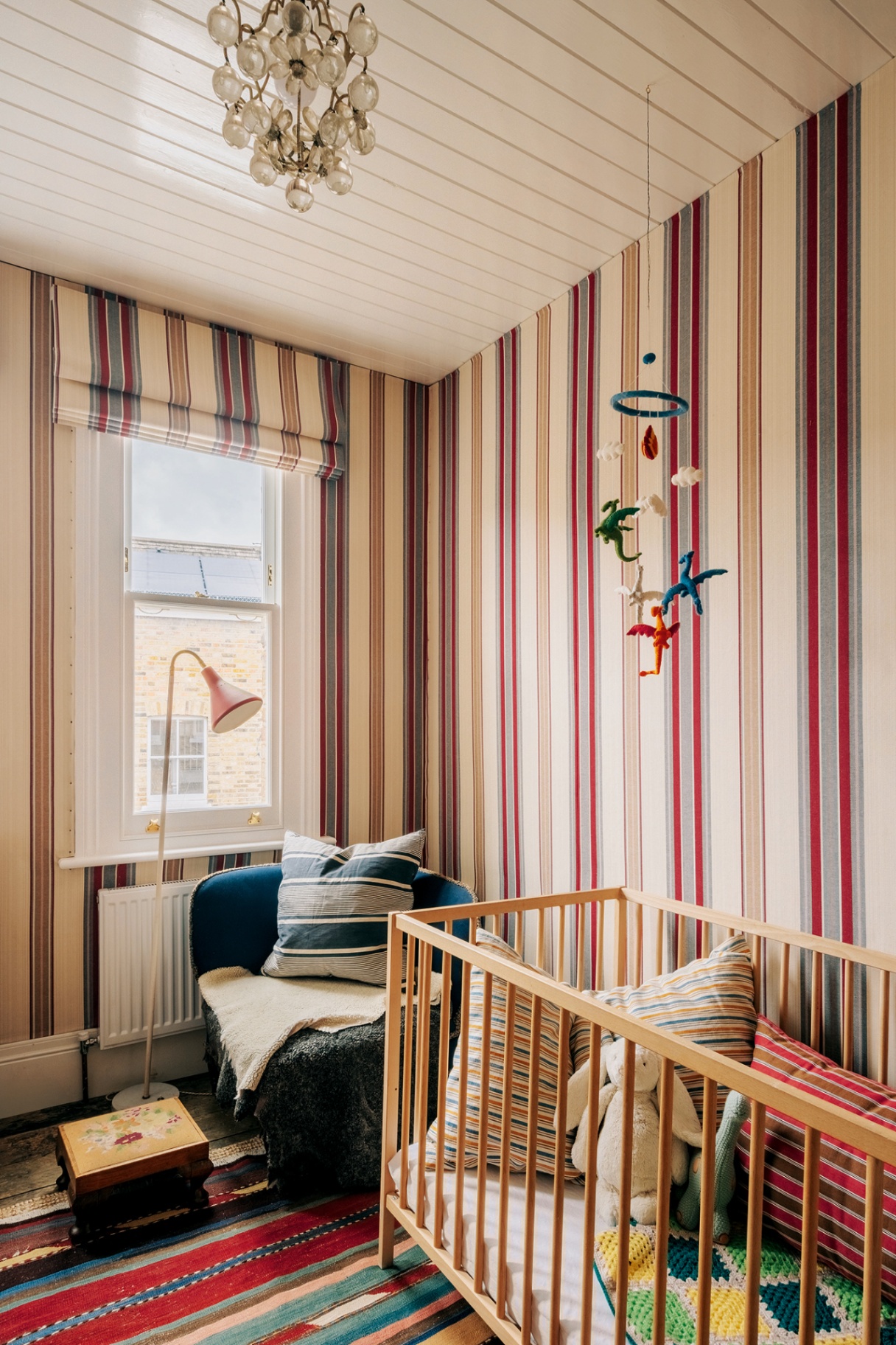 a nursery with striped walls, rug and blind, with a wooden cot filled with cushions and stuffed toys, an accent chair topped with a cushion and blanket, a floor lamp and an animal-themed mobile.