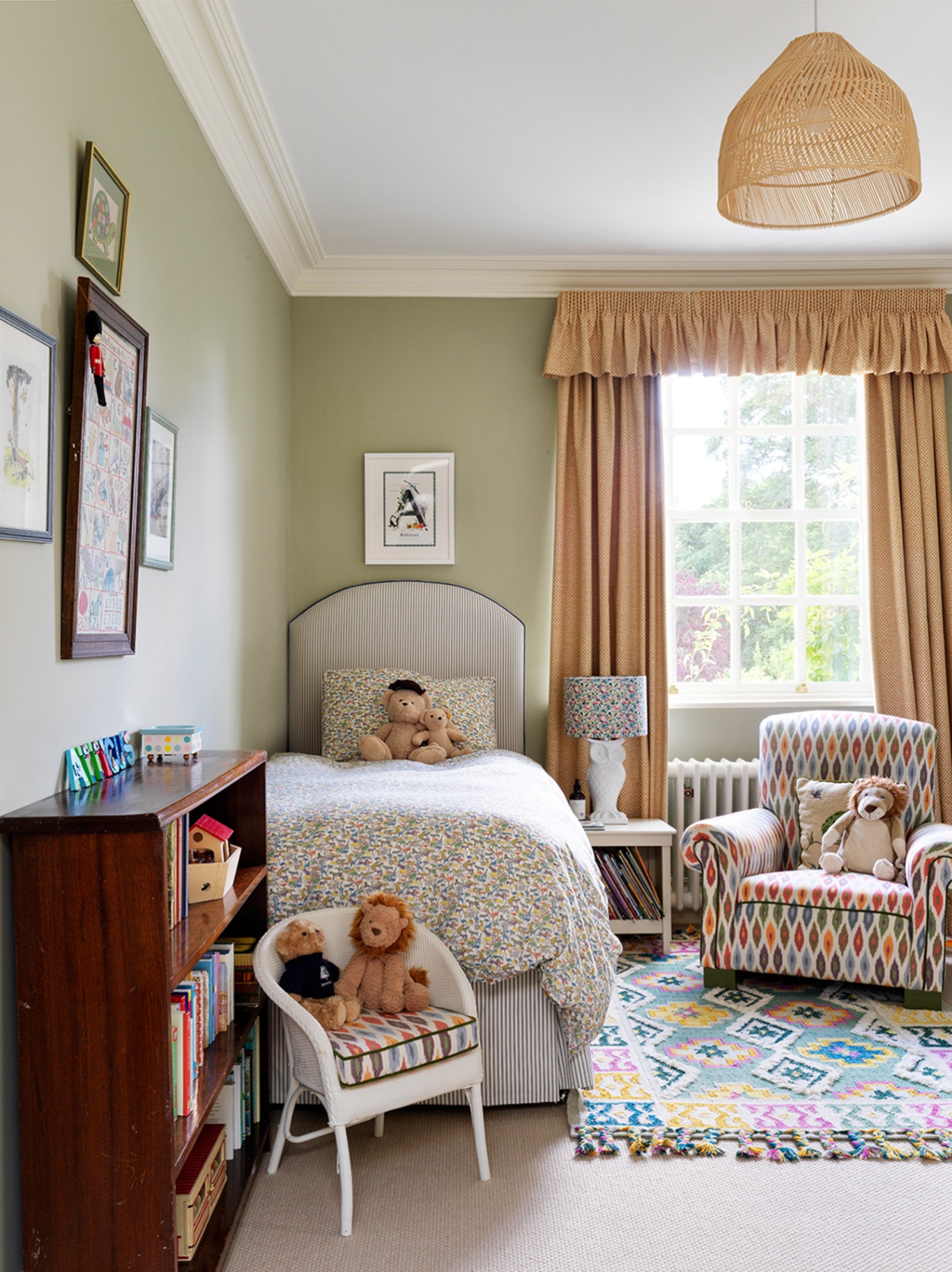 A light green hildren’s room with a patterned armchair, tasselled colourful rug, single bed topped with floral bedding and a teddy bear, a mini chair with stuffed toys on top and a wooden bookshelf