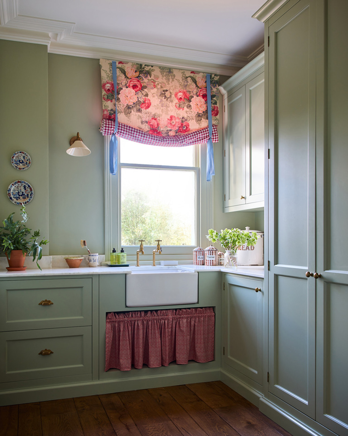 a utility room with sage green cabinetry, a dark wood floor, a window with a rose and gingham patterned blind, a belfast sink and a red patterned skirt under it
