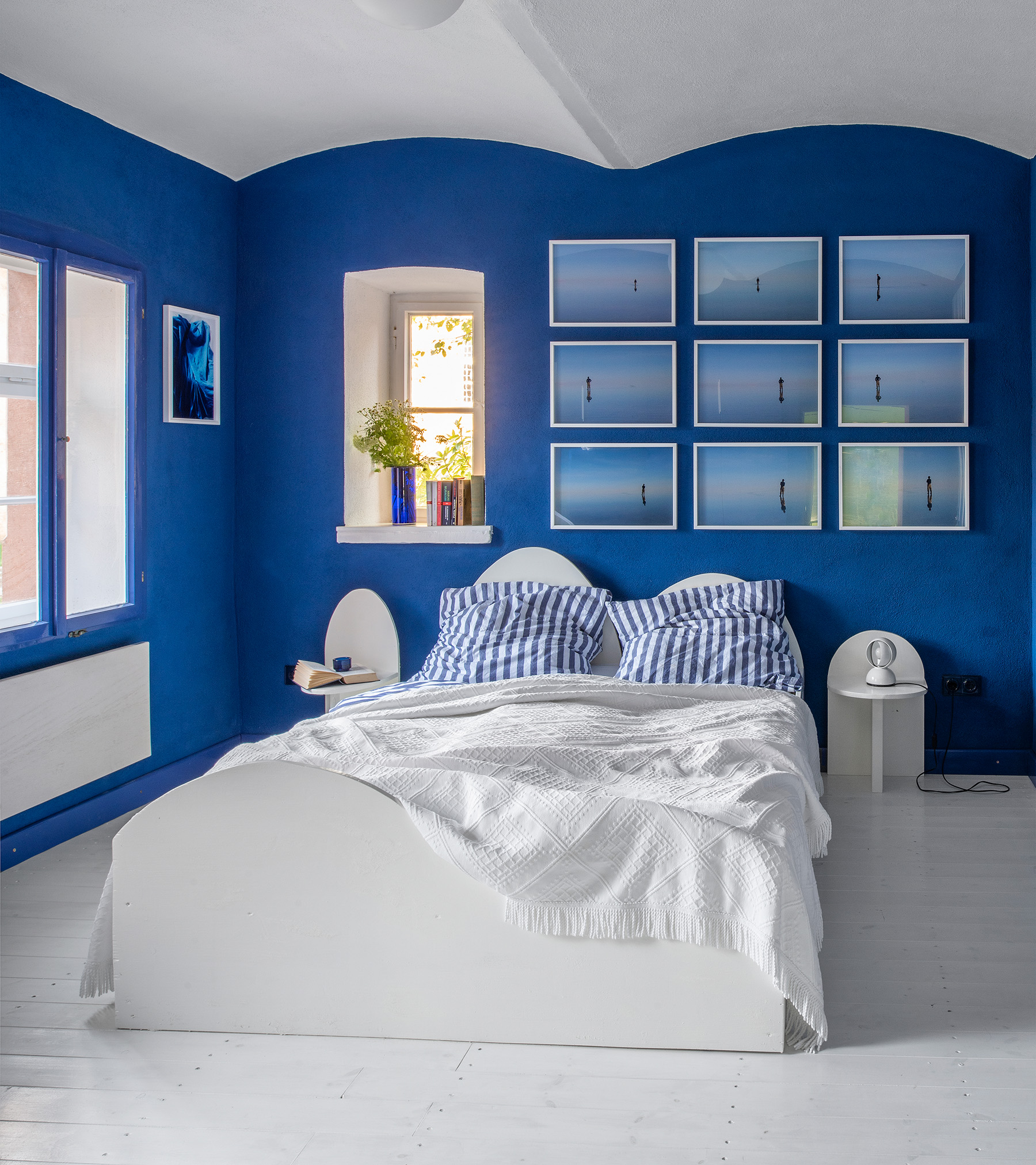 a bedroom with bold blue painted walls with a white frame around the ceiling, a white floor and a double bed with white bedding and blue and white stripe pillows