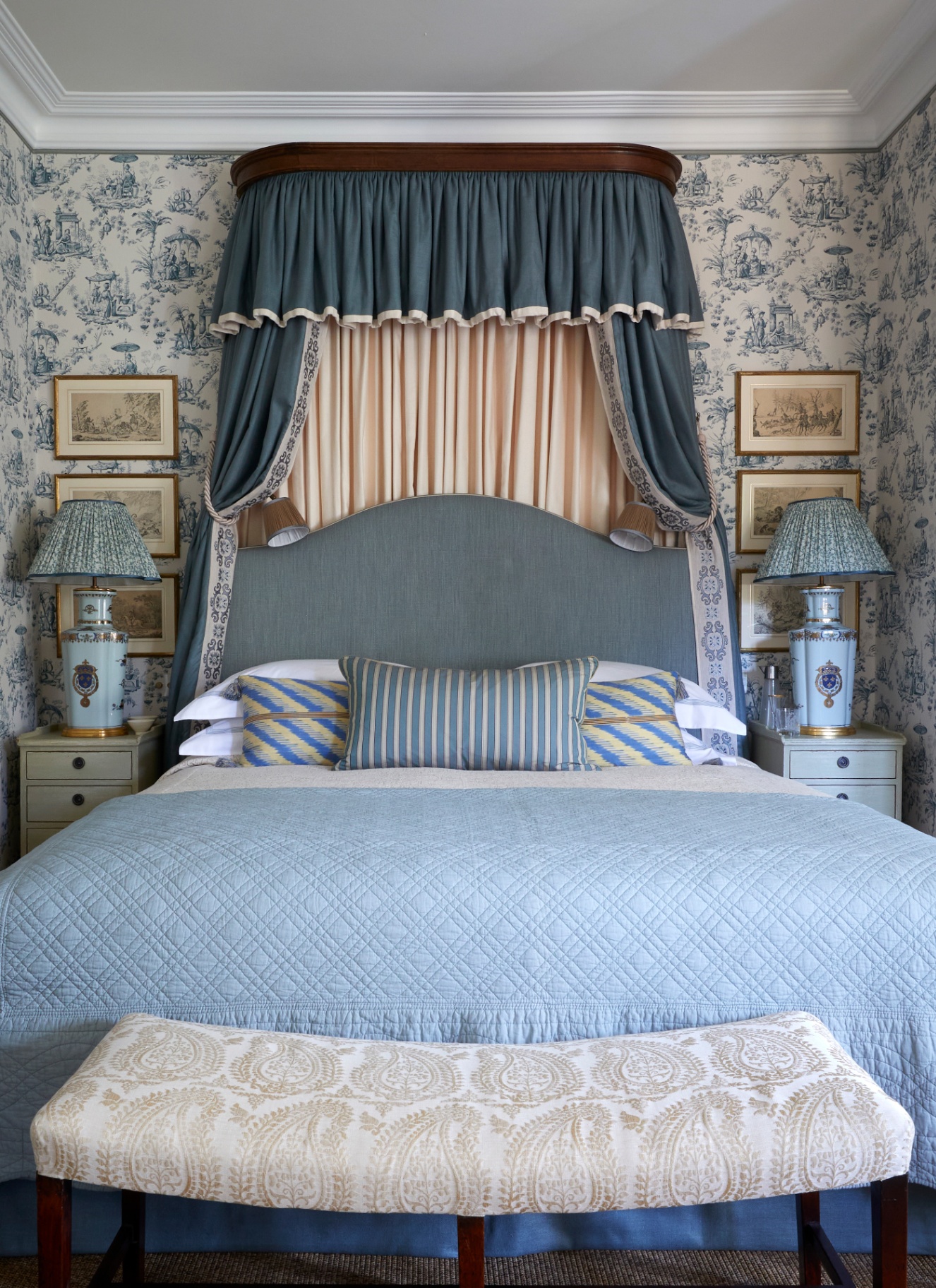 a bedroom with blue and white patterned wallpaper, a large bed with a blue canopy and a blue bedspread and a white and cream paisley patterned upholstered bench