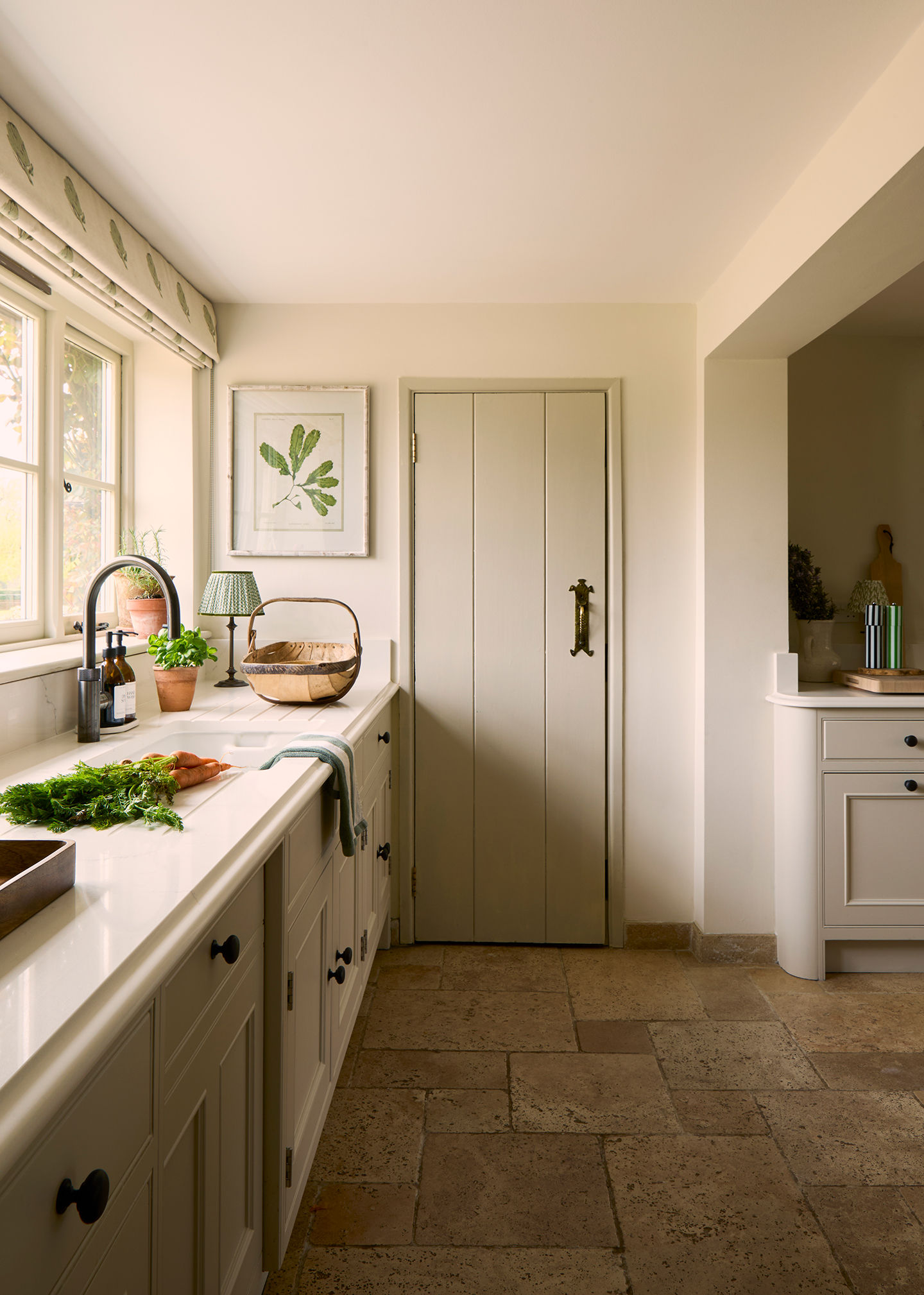 A close up of a tongue-and-groove door in a neutral kitchen with cabinets running along the left side and flagstone flooring. A sink sits below a window which is dressed with a blind made in an artichoke-print fabric