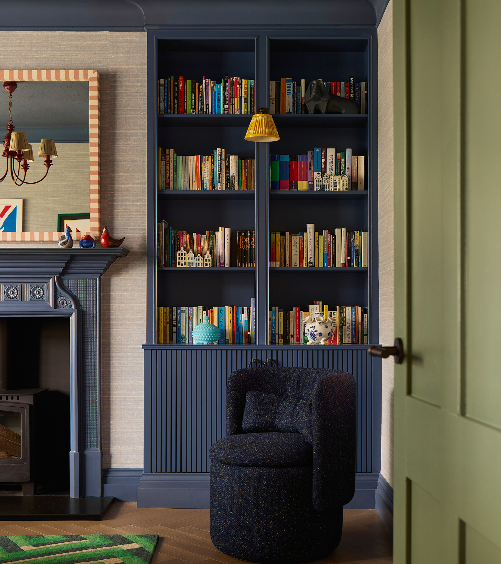a living room corner with a dark blue painted alcove filled with colourful books, a blue fireplace to the left and a small dark navy tub chair in front of the alcove