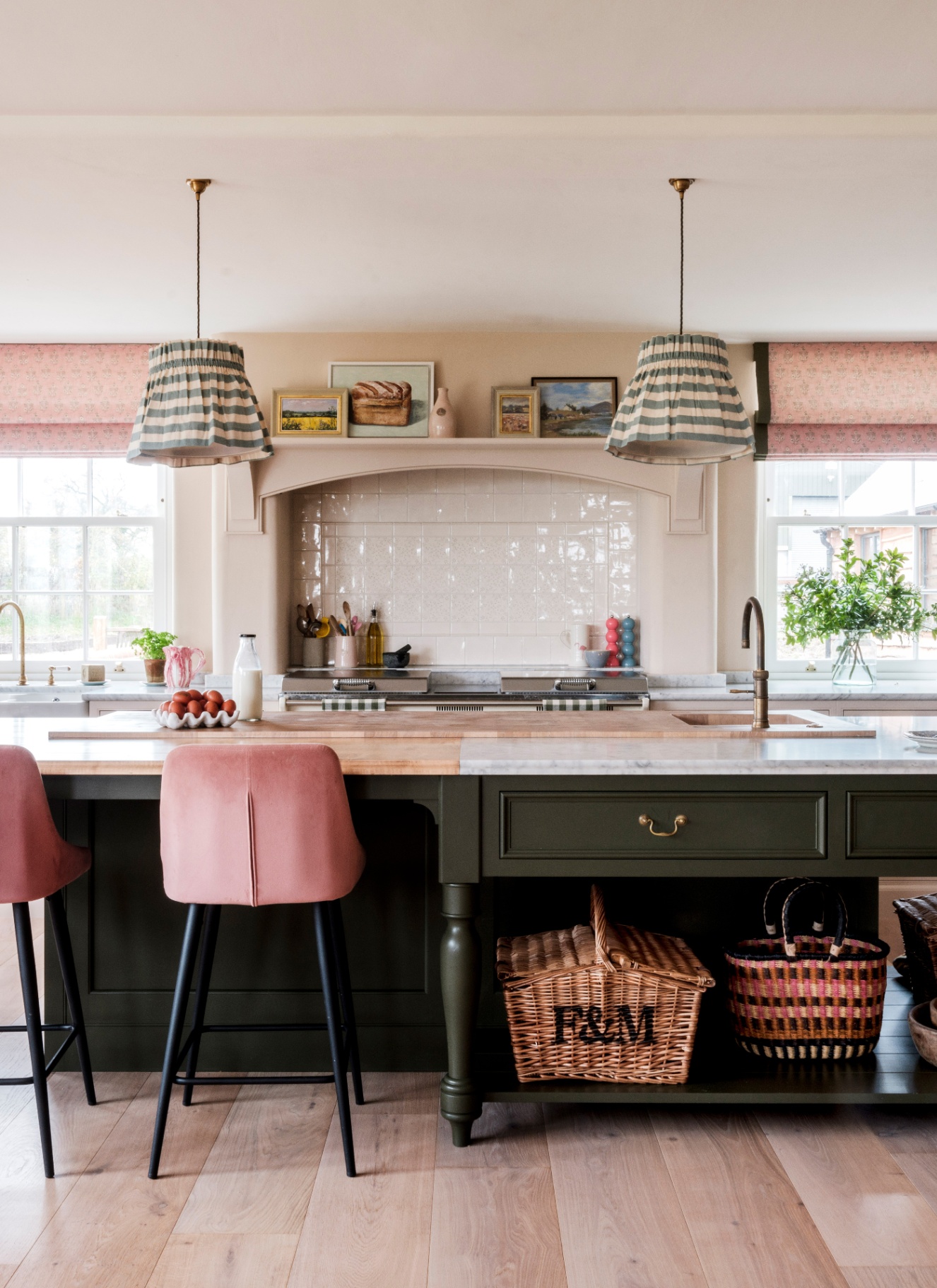 a kitchen with a large dark green kitchen island with half the top in wood and half in marble, with two striped pleated pendants above it and wicker baskets under neath it and two pink velvet stools