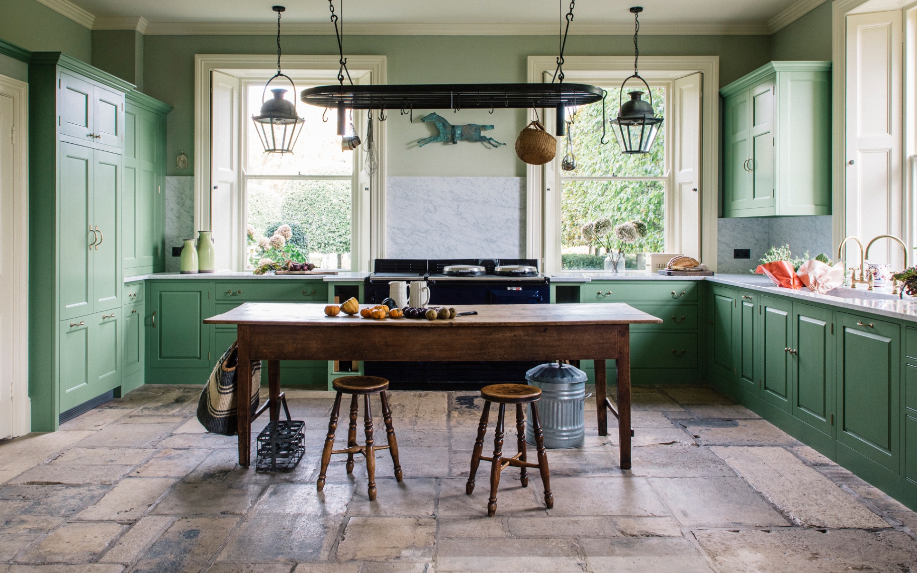 a country kitchen with a large wooden farmhouse table in the centre, bright green cabinetry, flagstone flooring and a large handing rack hanging above the table