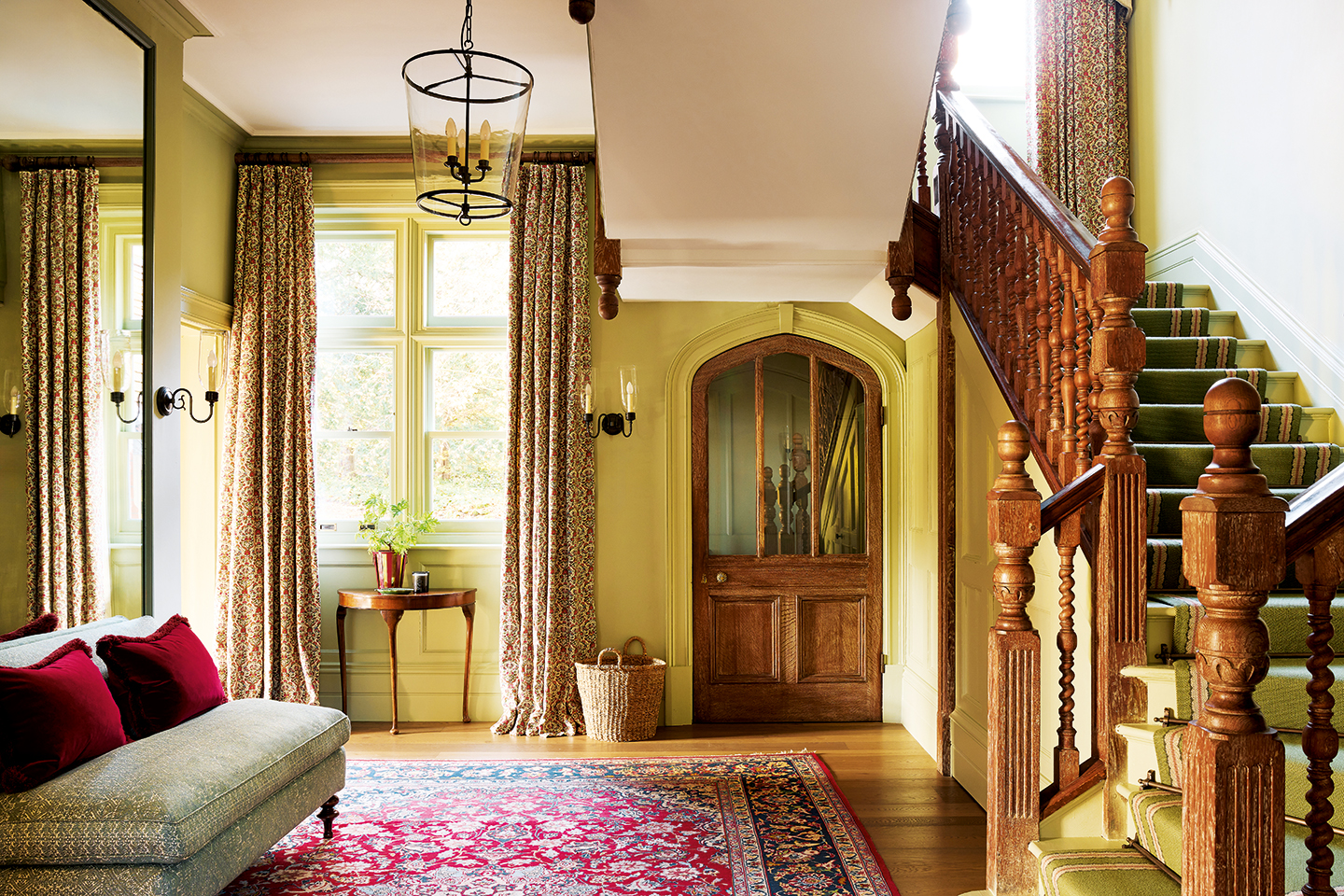 A grand entrance hall in a country home. To the right is a wooden staircase and in the centre is a large wooden door with glass panelling. In the foreground is a patterned red and blue rug and an upholstered bench to the left. A glass lantern hangs above