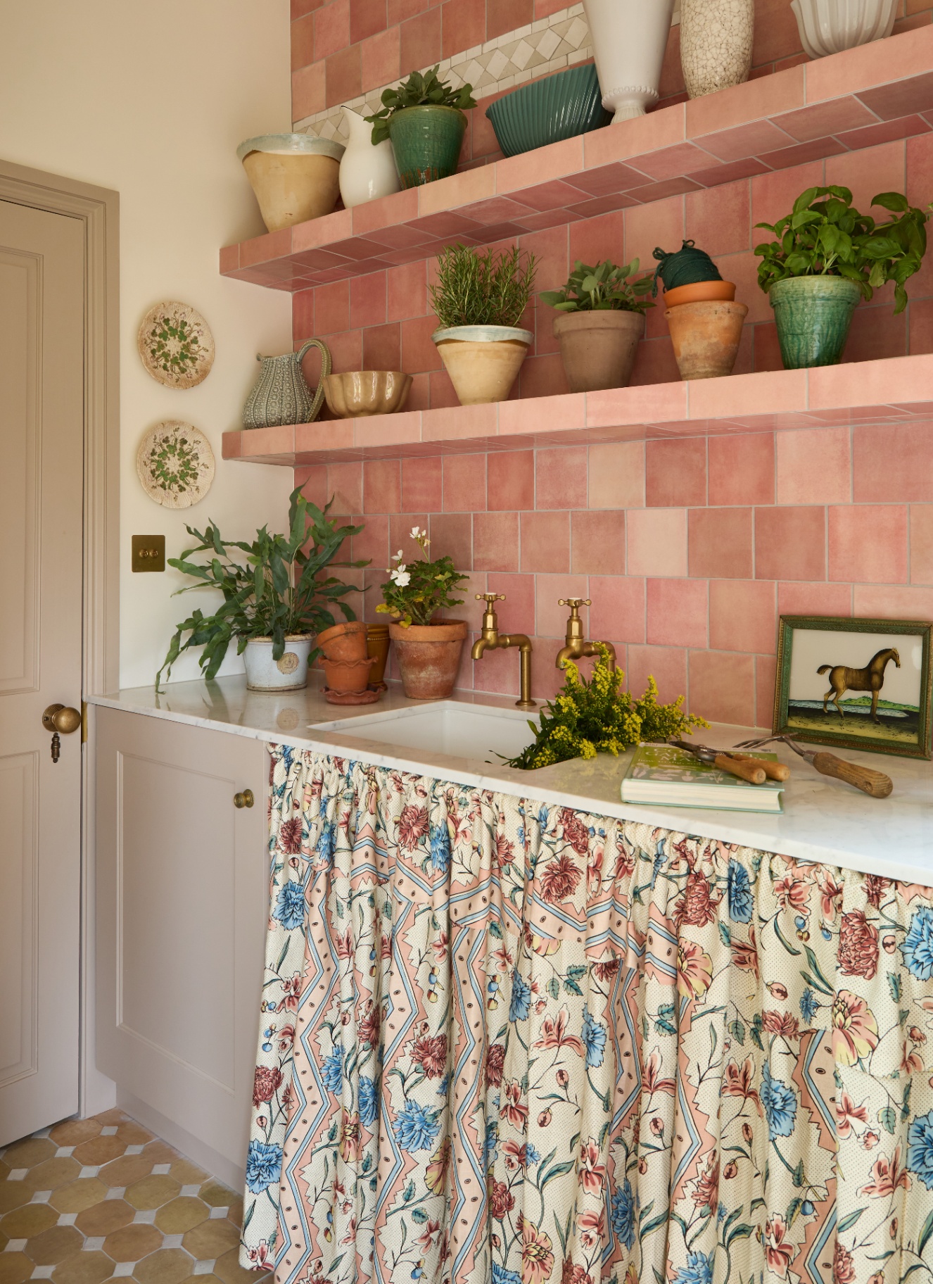 a utility room with pink zellige tiled walls and shelves, beige cabinets and a cream, pink and blue floral patterned sink skirt