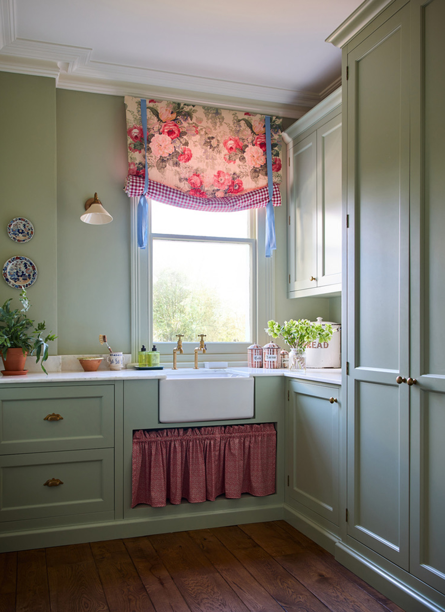 A sage green kitchen with a red patterned curatin under the sink and a floral and gingham blind on the window