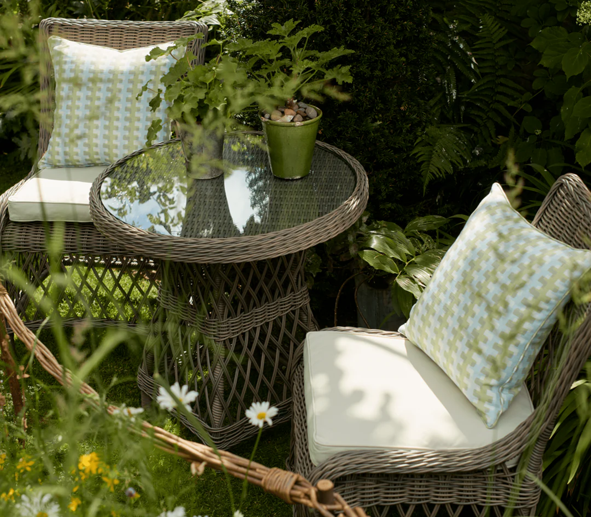 a close up of a wicker bistro set surrounded by greenery and daisies with neutral cushions and seat pads and plant pots on the table
