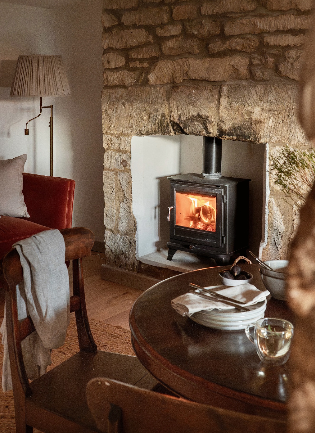 a living and dining area with a red velvet sofa, a wooden dining table and a large brick fireplace with a black wood burner