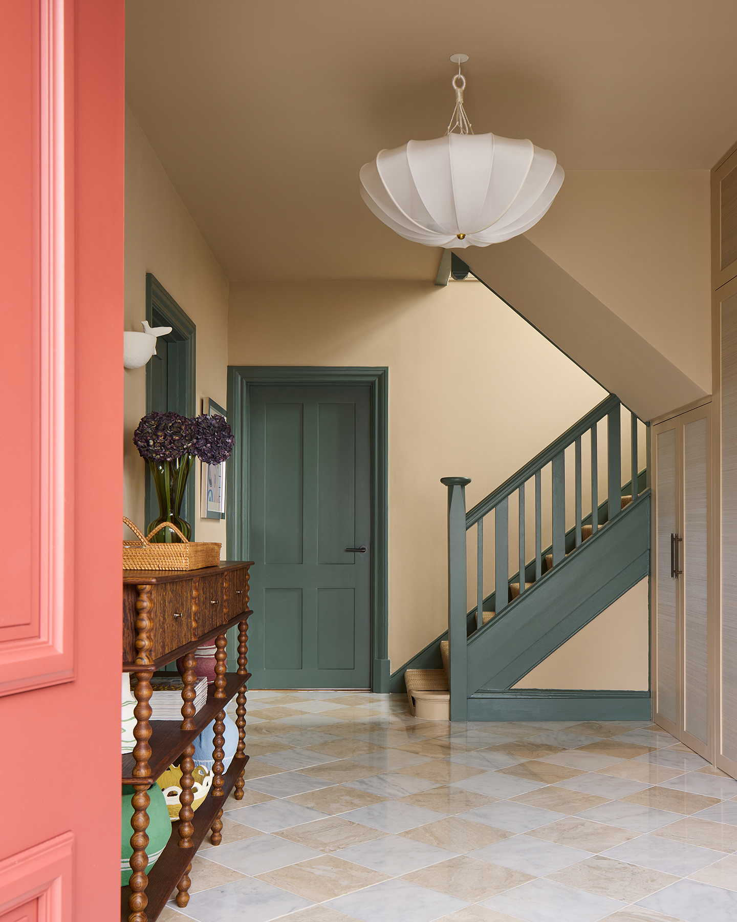 An entrance hall with a slither of a pink front door showing on the left. The floor is checkerboard marble tiles and doors and stairs painted in a dark green can be seen in the background. A large pendant hangs and to the left is a wooden bobbin console