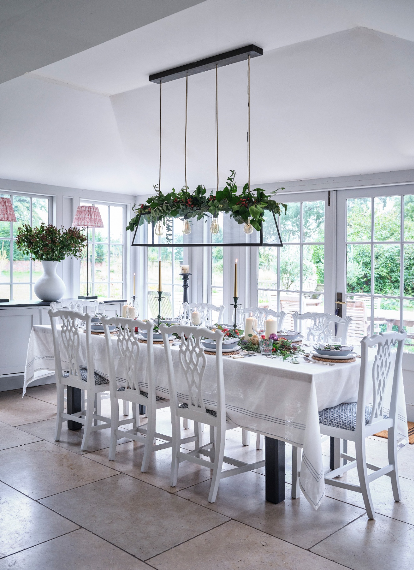 A kitchen island with a green pitcher of crimson amaryllis, potted hyacinths, fruit, and candles, with a sink, a lamp, and hanging aprons in the background.