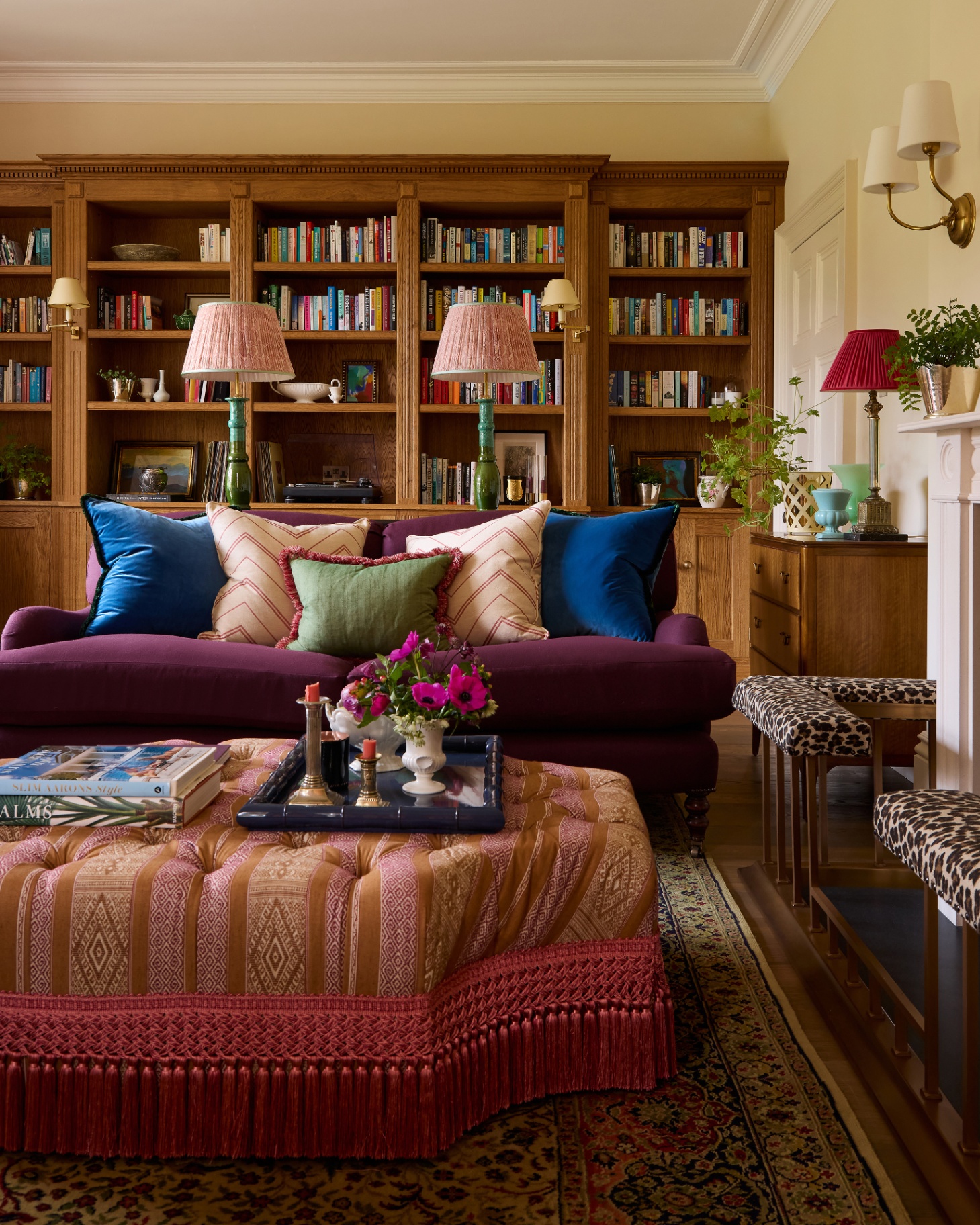 a living room with a large wooden bookcase behind a dark purple sofa and a beige and pink patterned upholstered ottoman with a navy tray on top