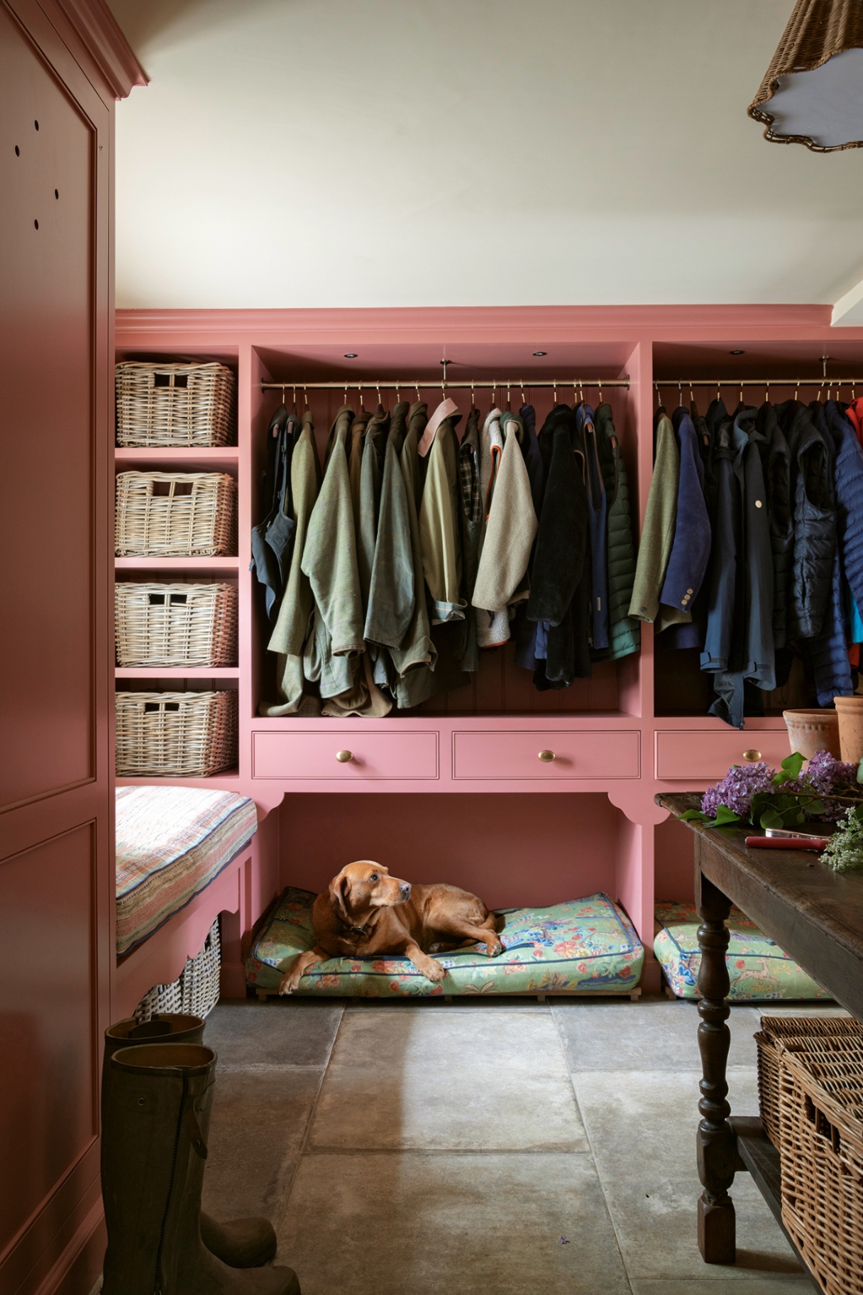 A boot room with pink painted joinery holding wicker baskets and a clothes rail filled with jackets. Flagstone flooring covers the room, topped with two colourful dog beds built into the joinery, one with a red labrador lounging on it