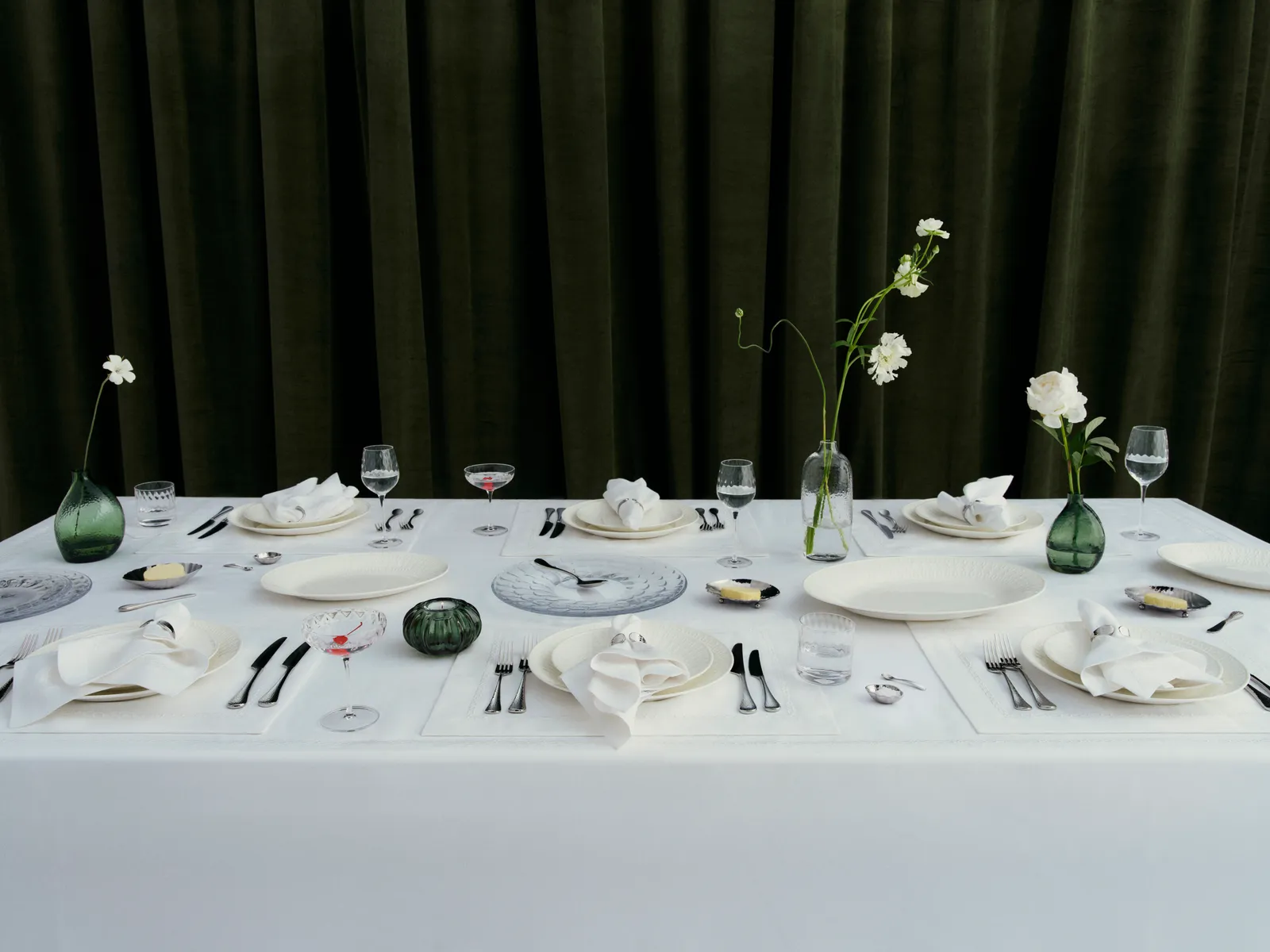 An elegantly set dining table with white linens, fine china, and glassware, accented by green glass vases with single white blooms against dark green velvet curtains