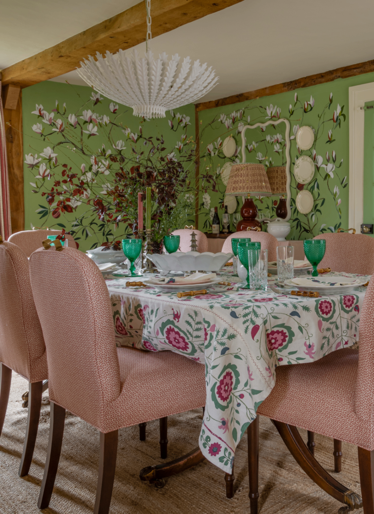 Country dining room with green magnolia wall mural, pink upholstered chairs, floral tablecloth, and a white palm chandelier
